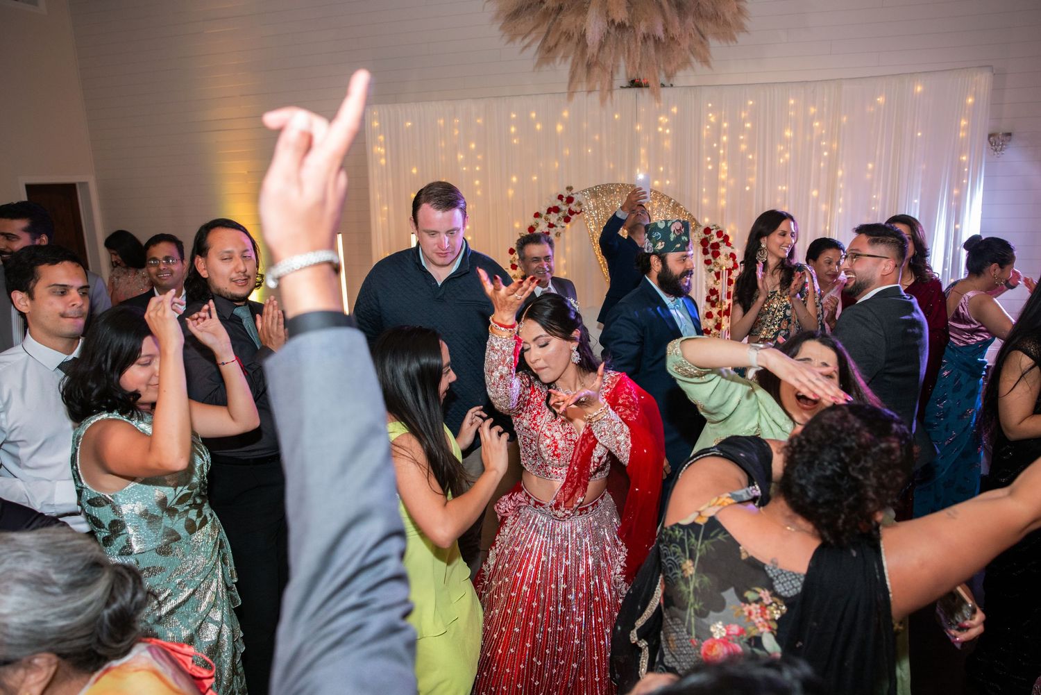 Guests in colorful formal wear dance joyfully at a lively Indian wedding reception celebration.