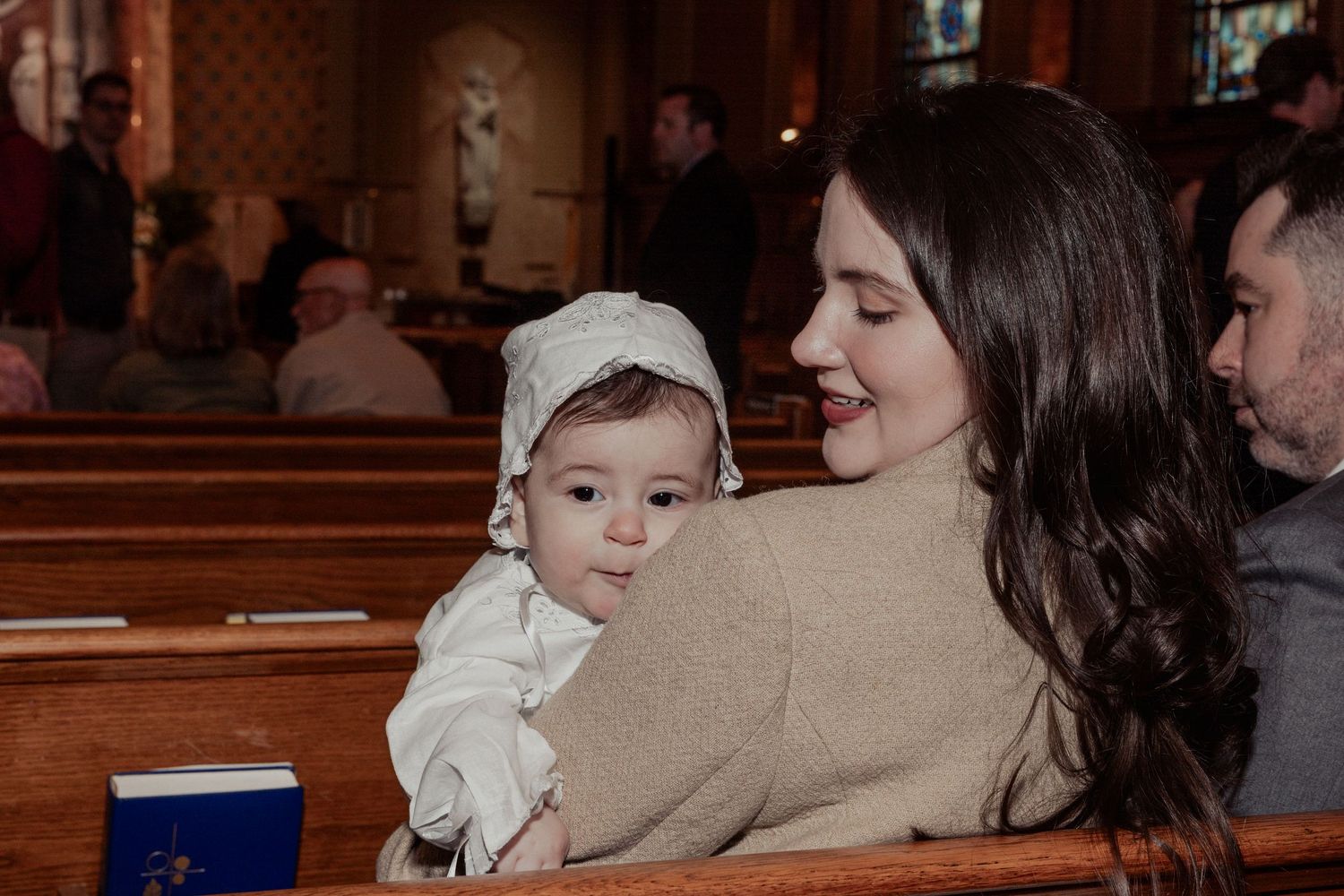 Young mother with dark hair smiles while holding her baby in a white christening outfit in a church pew.