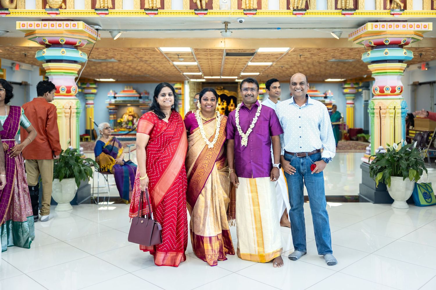 Four people in vibrant traditional Indian sarees and kurtas posing inside a decorated Hindu temple.