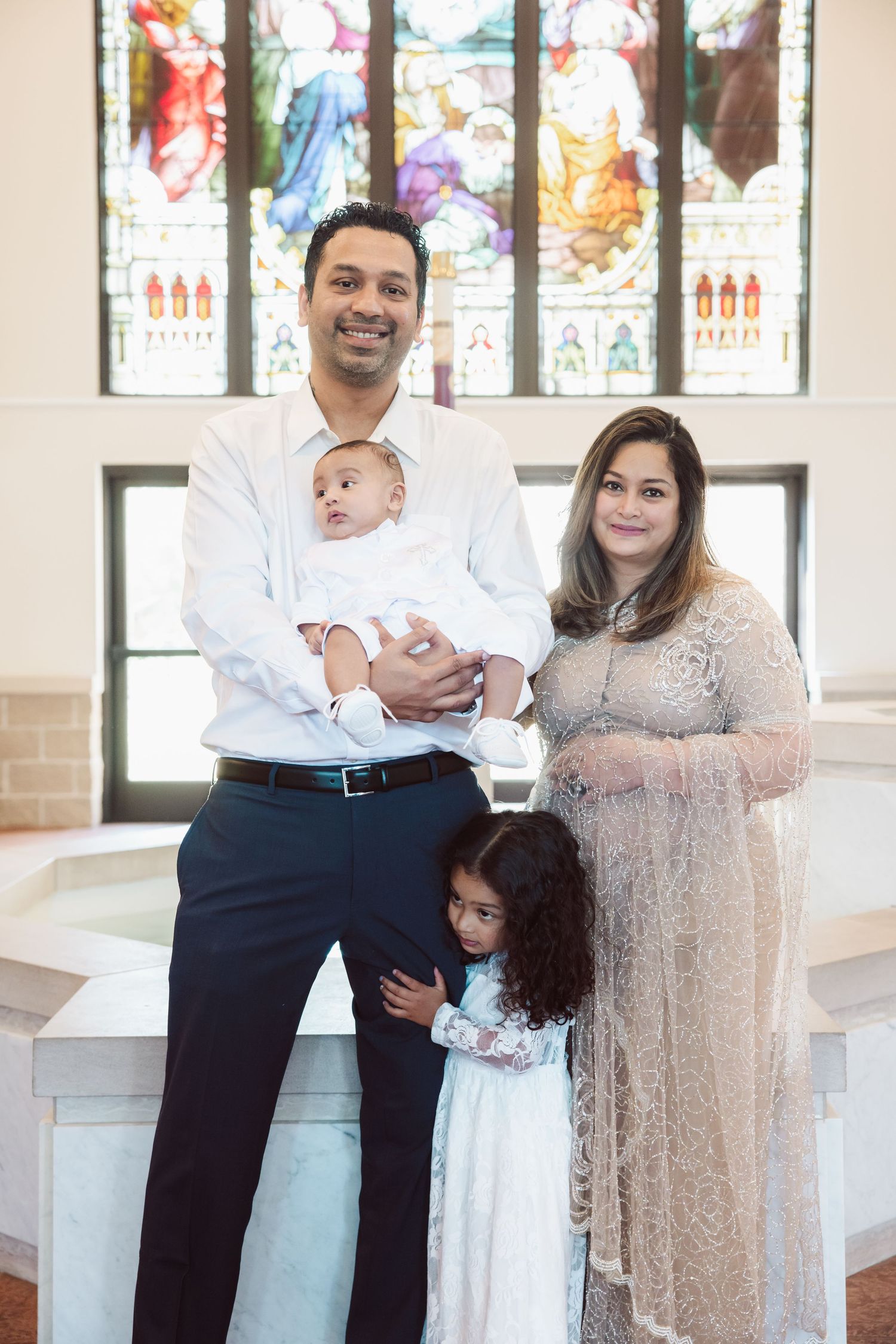Family posing with newborn baby and young daughter in front of stained glass church window.