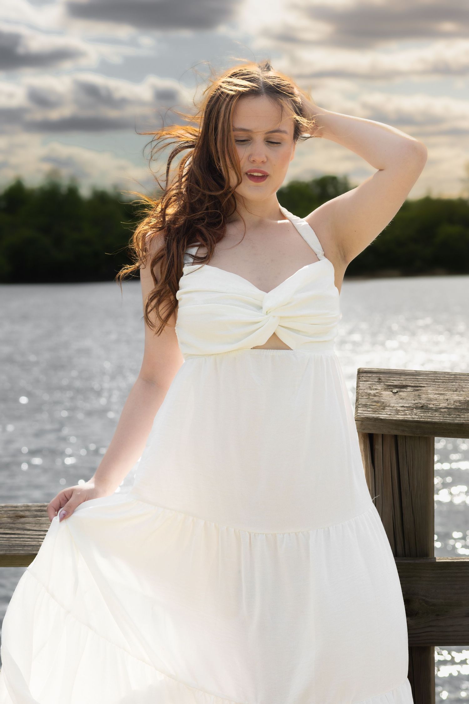 A woman in a flowing white sundress stands on a wooden dock by a lake, her hair blowing in the wind.