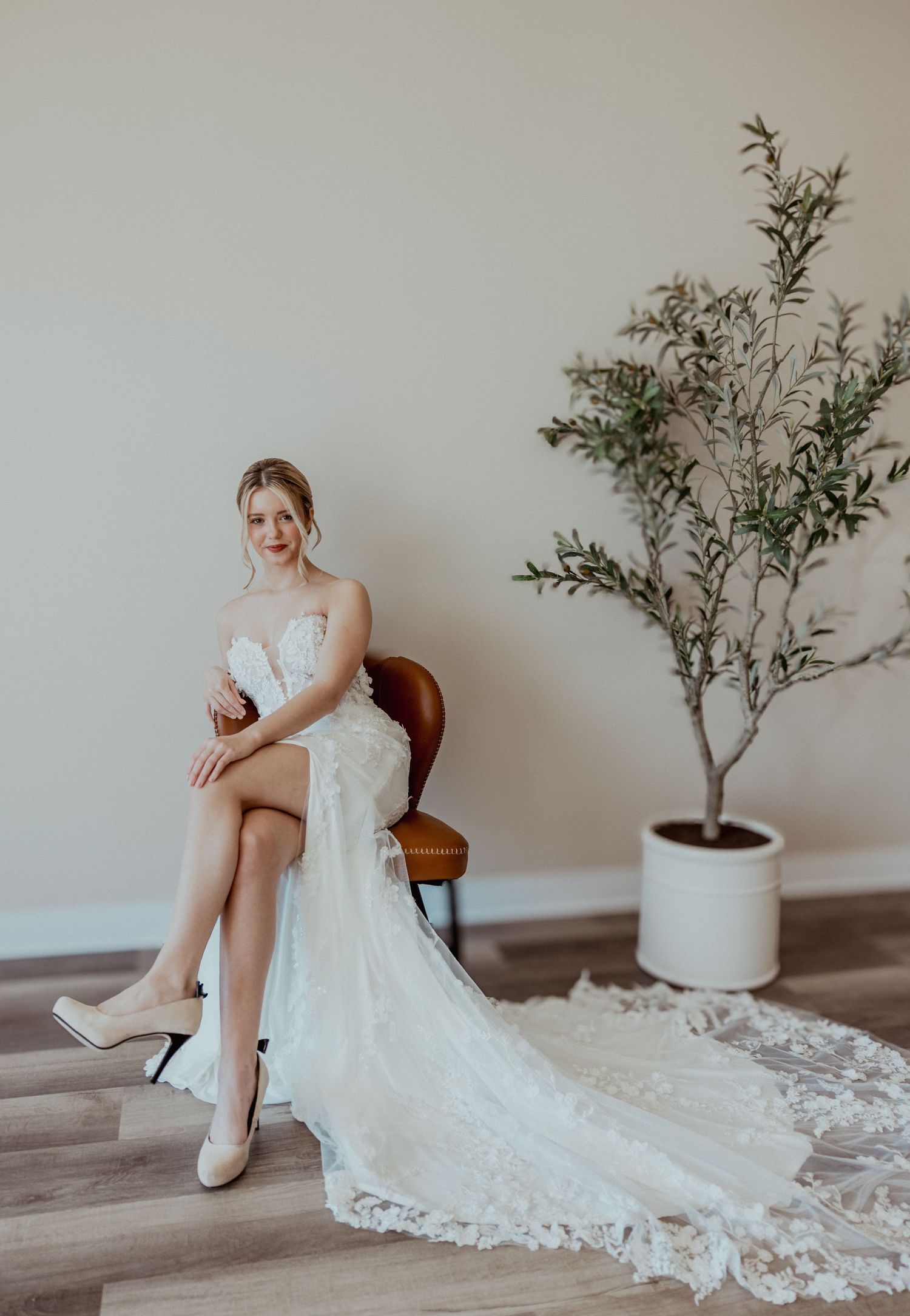 Elegant bride in lace wedding gown with train seated on chair next to potted olive tree.