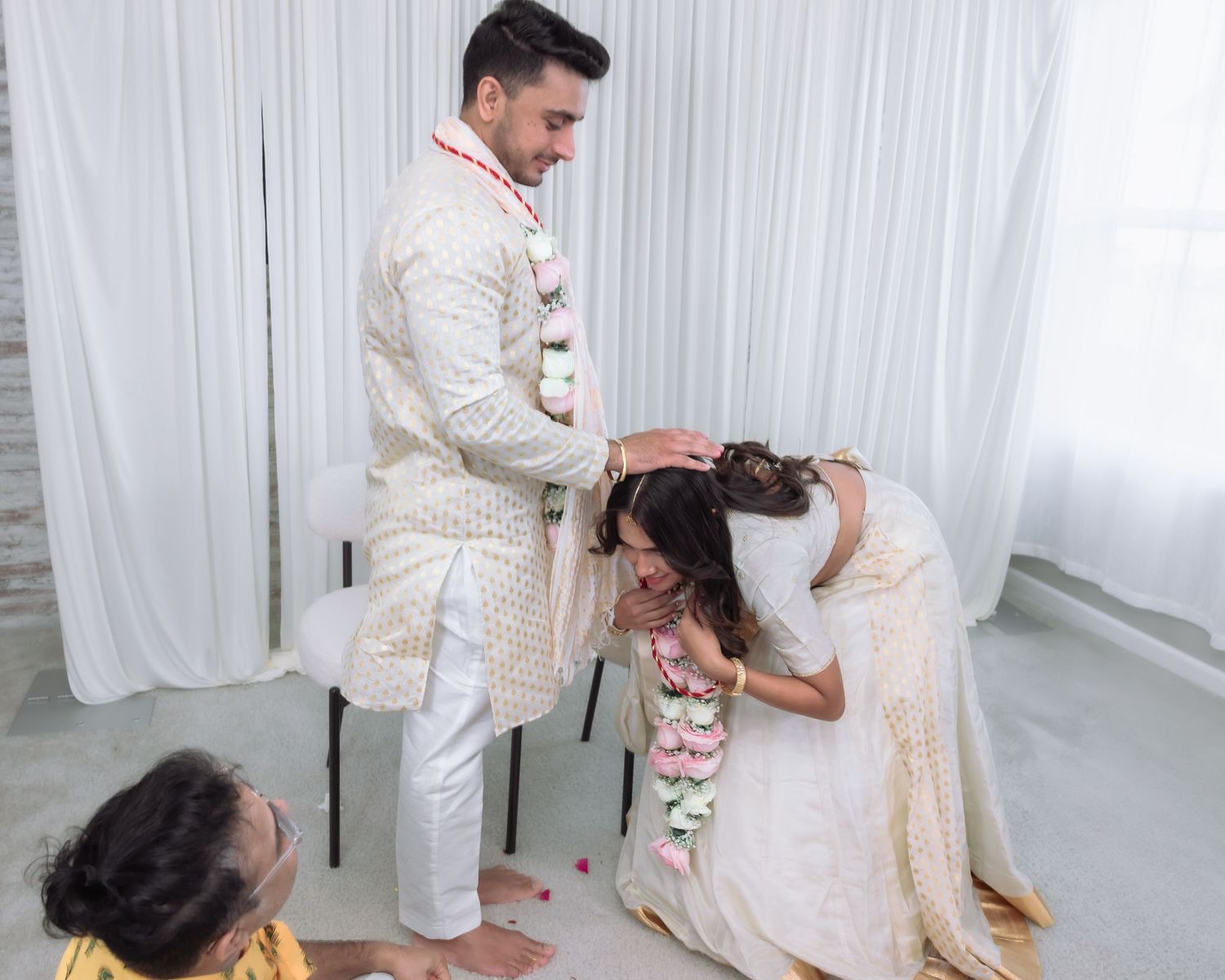 Indian groom blesses kneeling bride during traditional Hindu wedding ceremony with elder present.