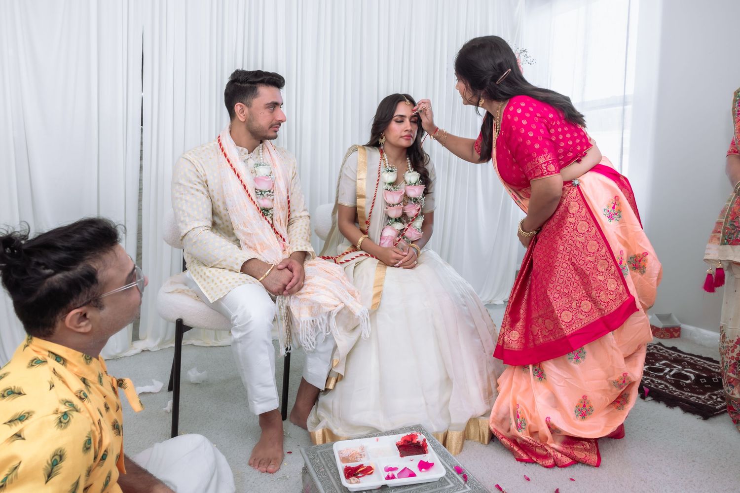 Woman in red saree applying tilak to Indian bride during traditional Hindu wedding ceremony with guests.