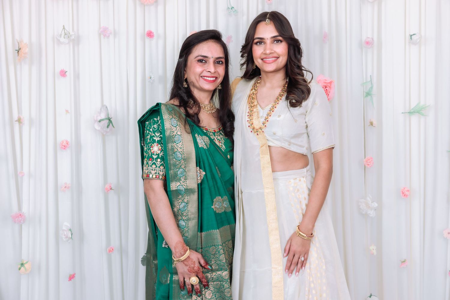 Mother and daughter posing at Indian wedding, wearing elegant green saree and white lehenga with gold jewelry.