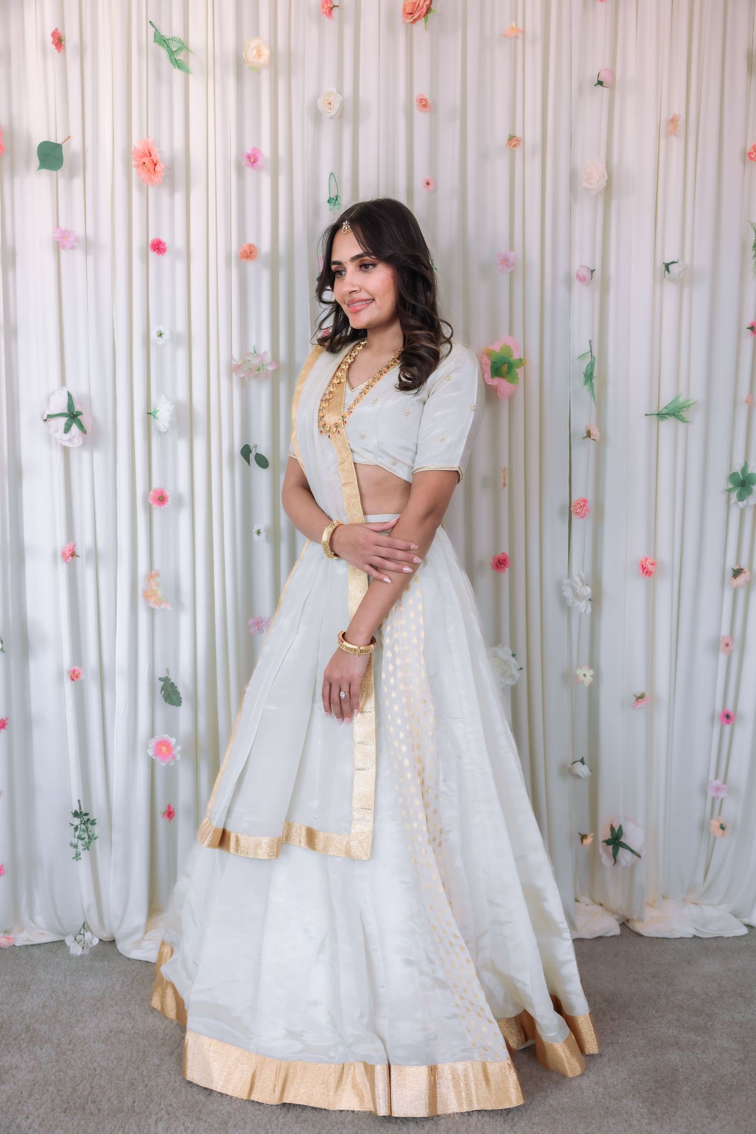 Smiling young woman in white gold-bordered lehenga standing before a pastel floral backdrop.