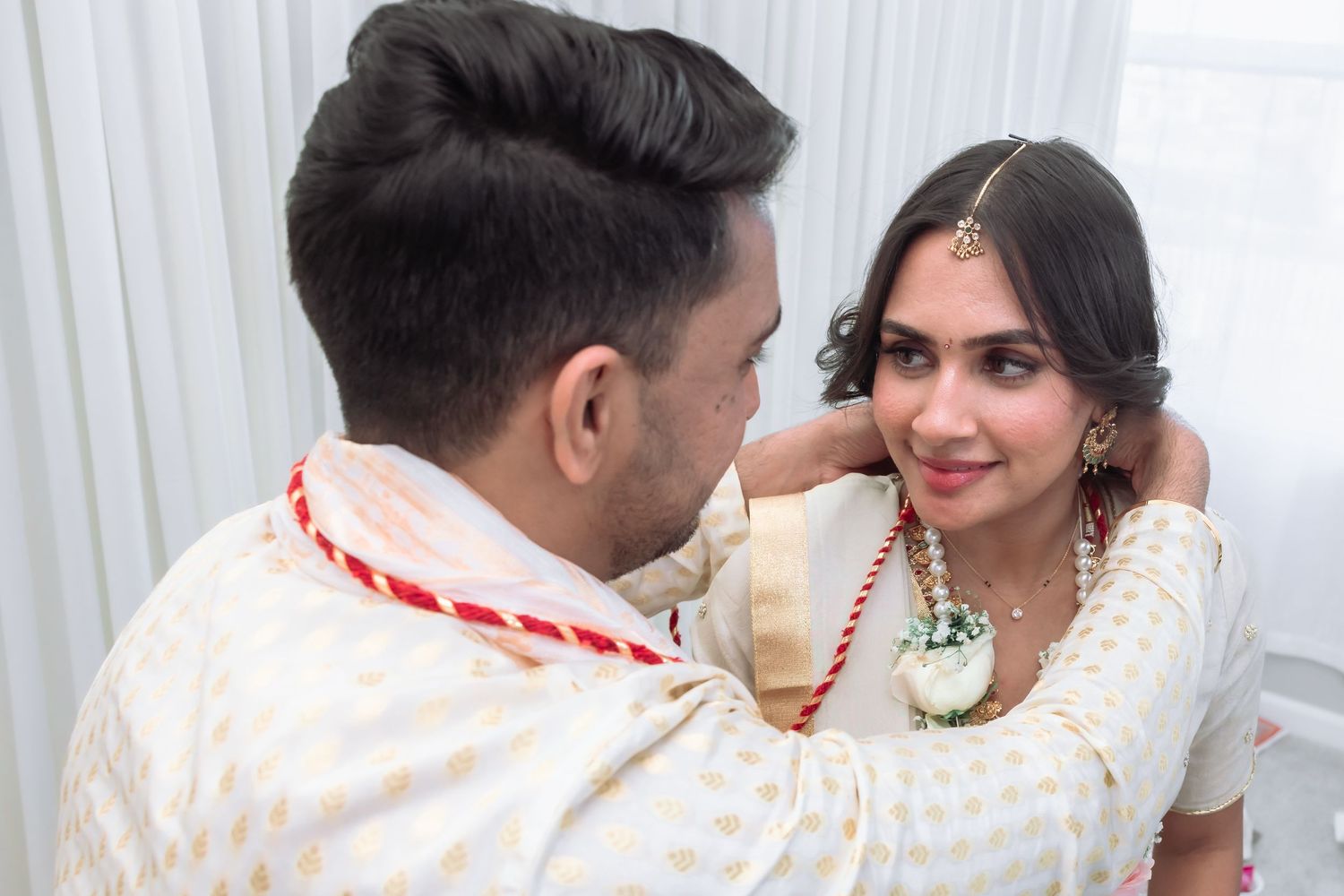 Groom adjusts bride's necklace during intimate Hindu wedding ceremony, bride smiles warmly.