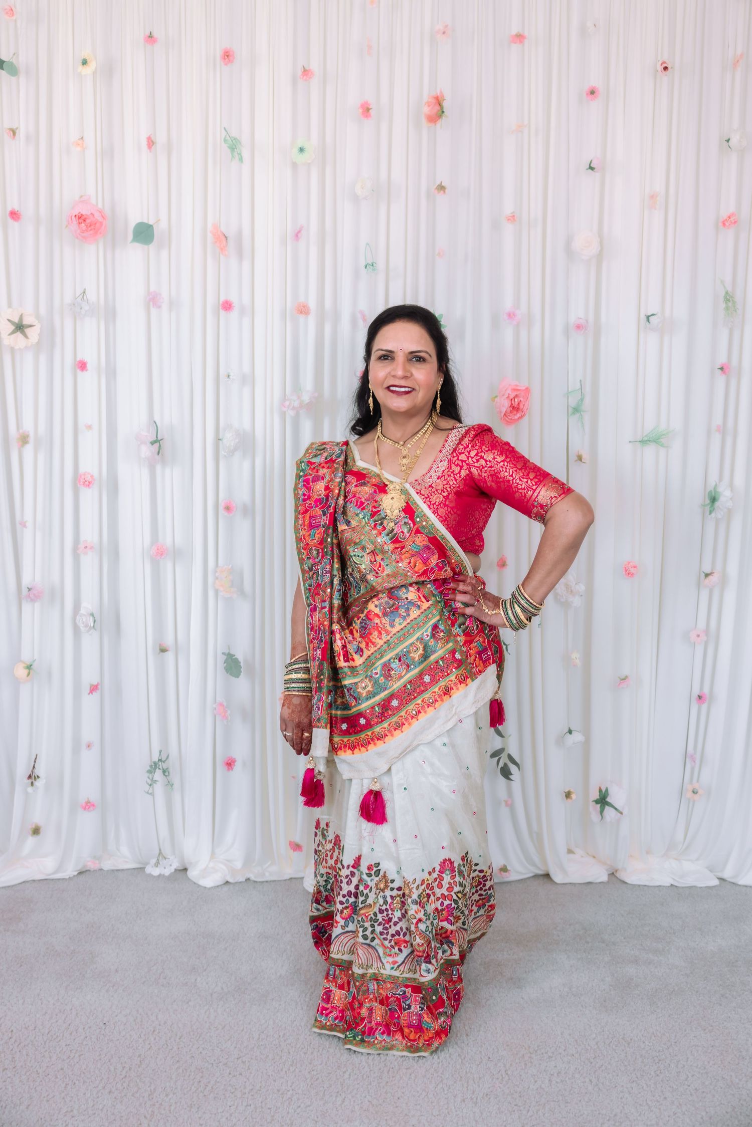 Indian woman in ornate white and red embroidered lehenga with jewelry poses at floral decorated wedding backdrop.