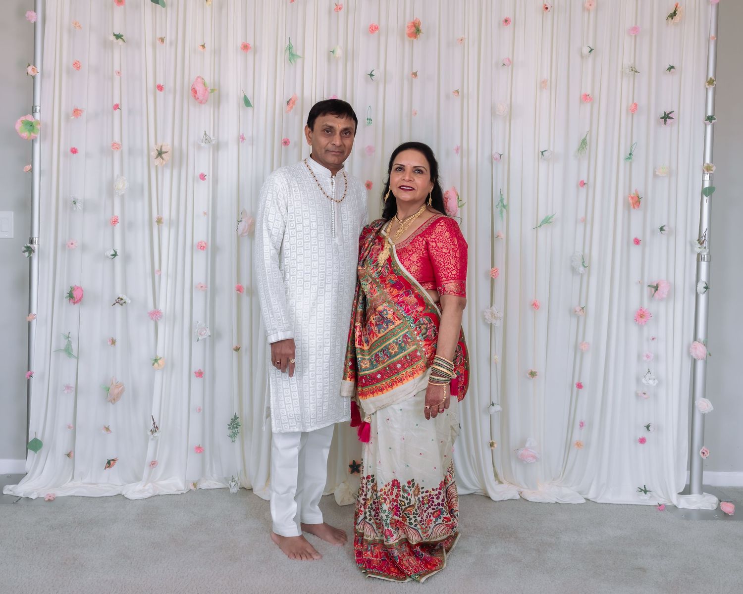 Indian couple in traditional attire posing together against a white floral backdrop at a celebration.