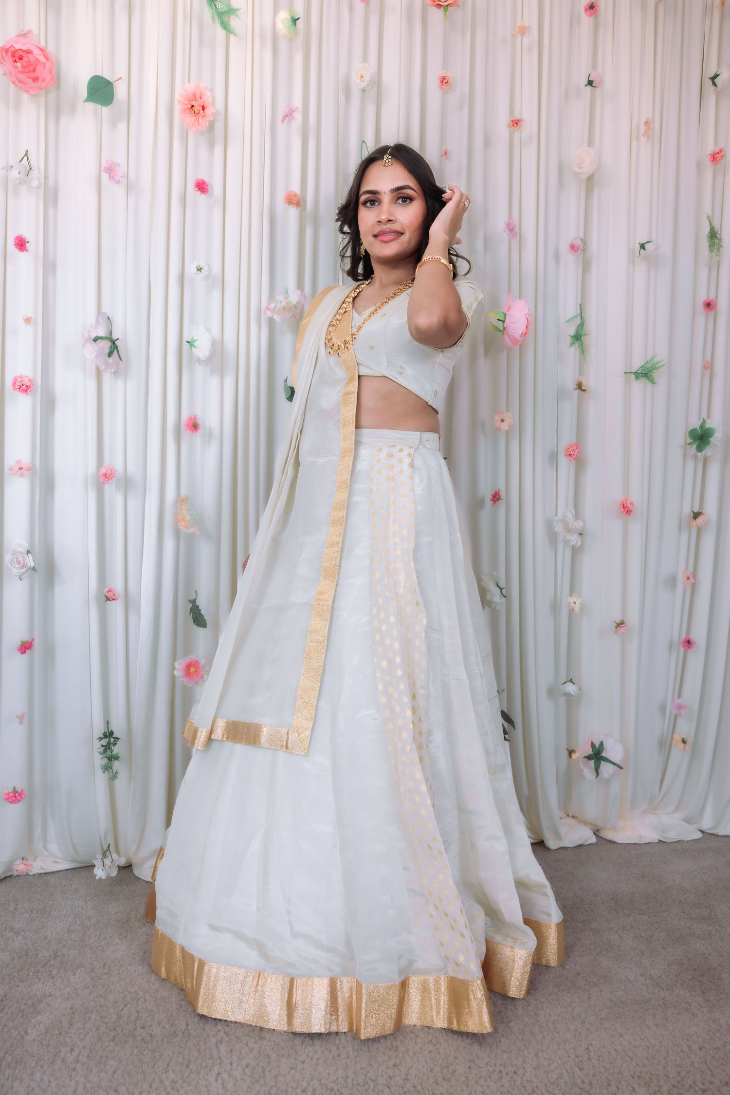 Indian bride in elegant white gold-trimmed lehenga extends hand toward camera at decorated backdrop.