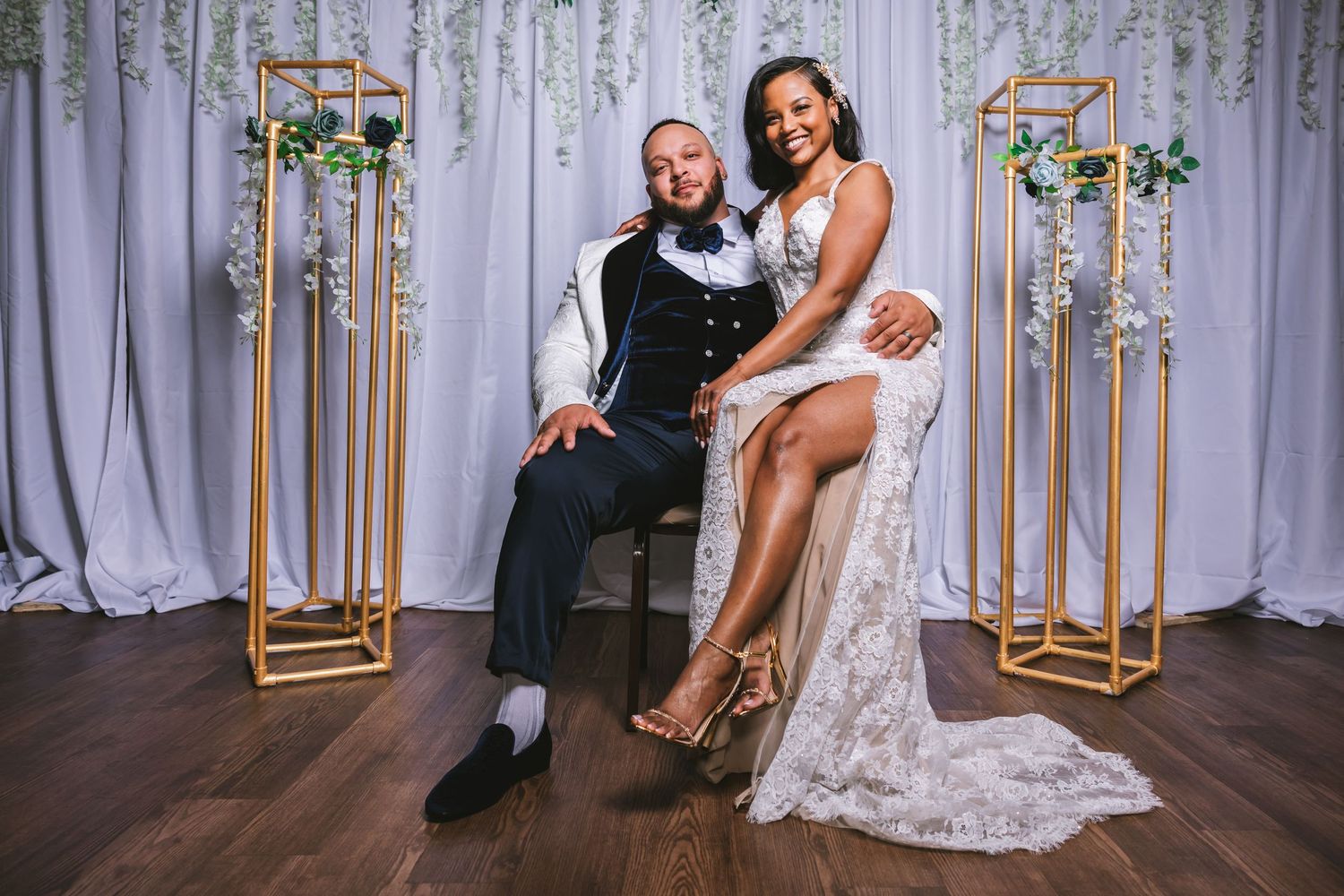 Bride in lace gown and groom in navy suit posing at elegant white and gold wedding backdrop.