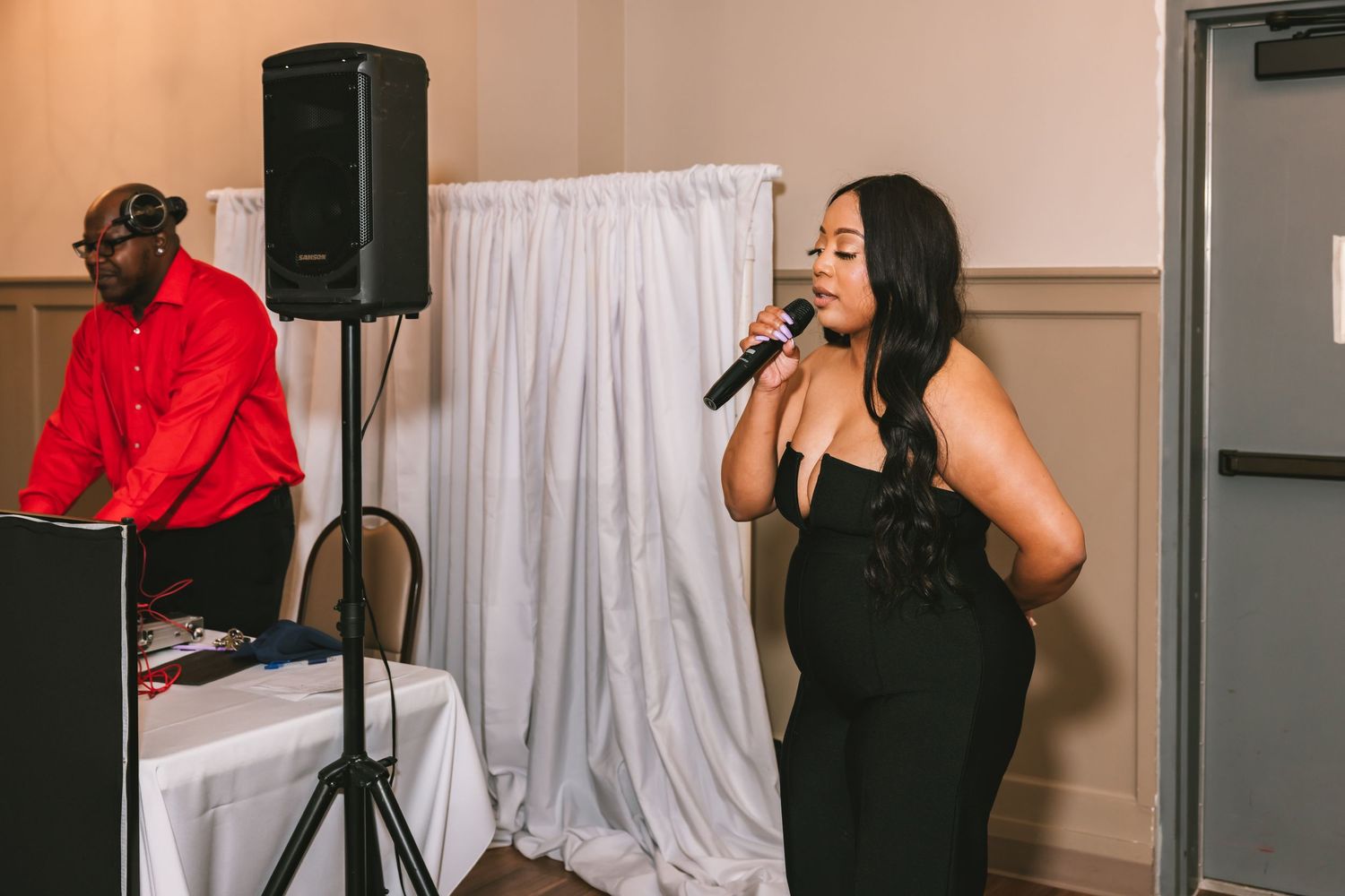 A woman in a black dress sings into a microphone at an elegant wedding reception with a DJ in the background.