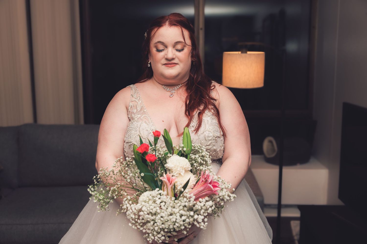 Plus-size bride in beaded lace wedding dress admires bouquet of pink lilies and red tulips.