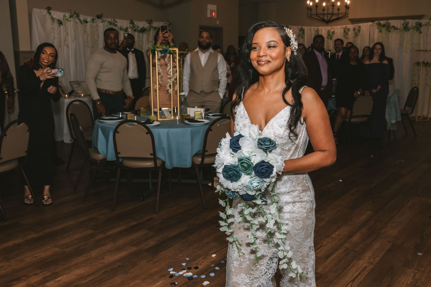 Radiant bride in lace gown holds navy blue and white bouquet while walking through wedding reception hall.