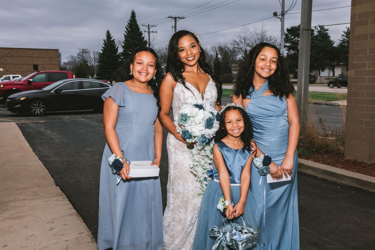 Bride in lace gown posing outdoors with three young girls in dusty blue bridesmaid dresses.