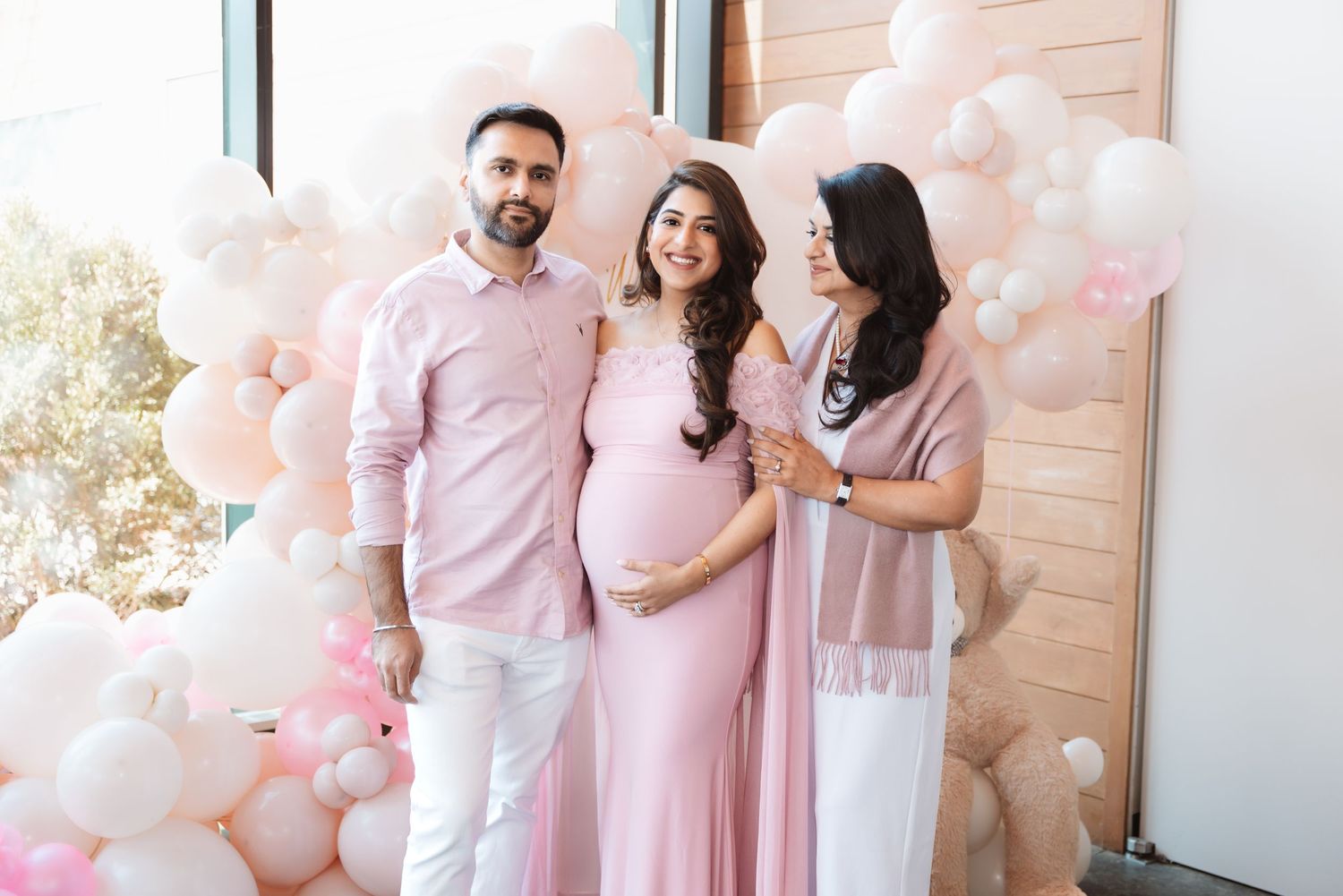 Pregnant woman in pink gown poses with man and woman in front of pink balloon arch at baby shower.