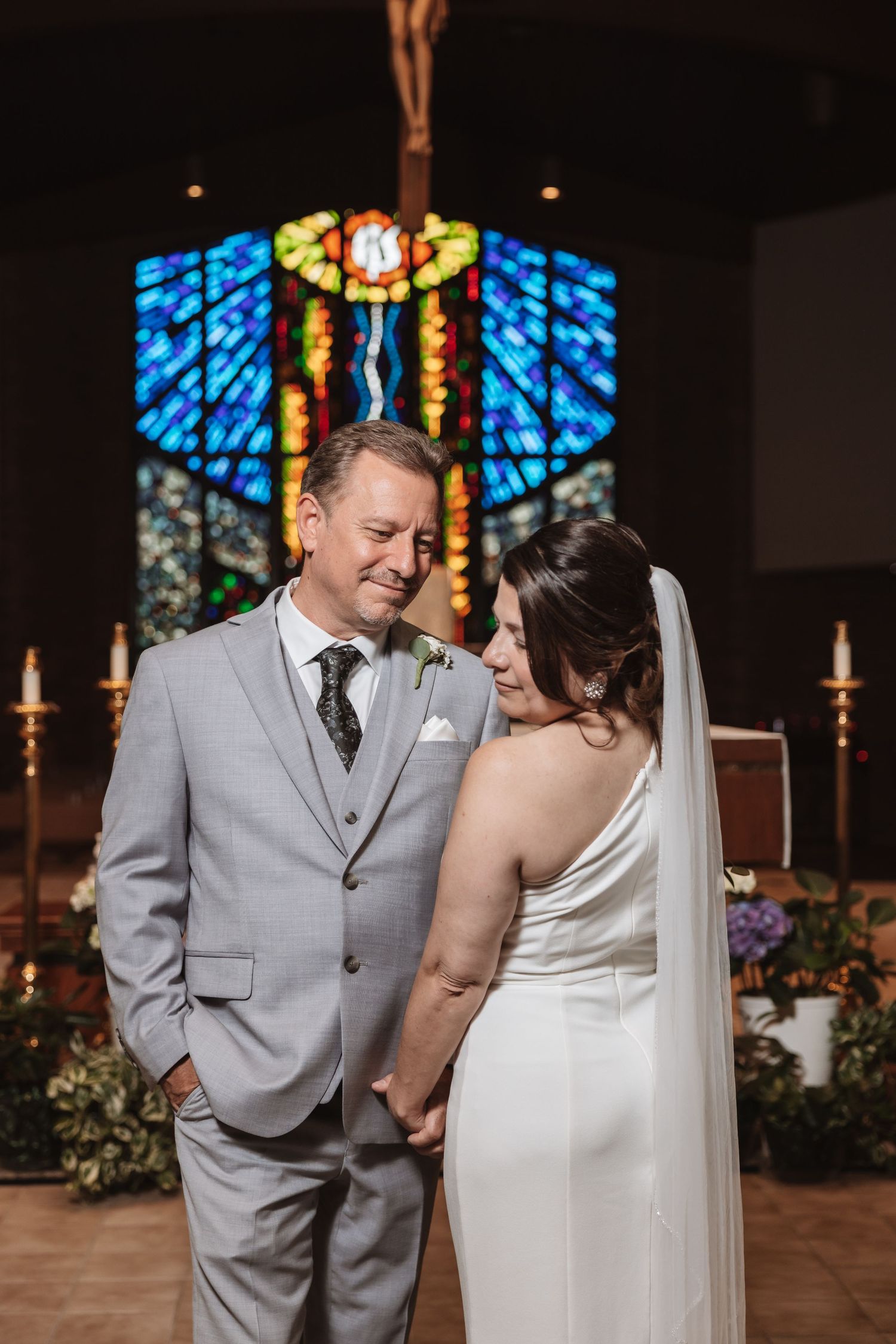 Bride and groom hold hands smiling at altar with colorful stained glass church window behind them.
