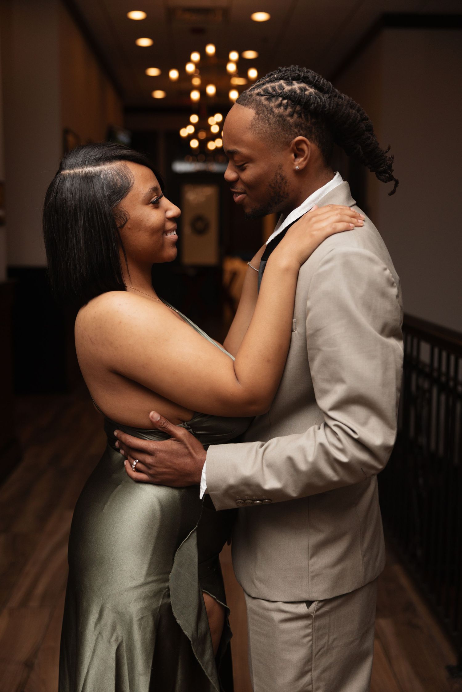 Elegant couple embracing and smiling in a warmly lit hallway, dressed in formal attire for a special occasion.