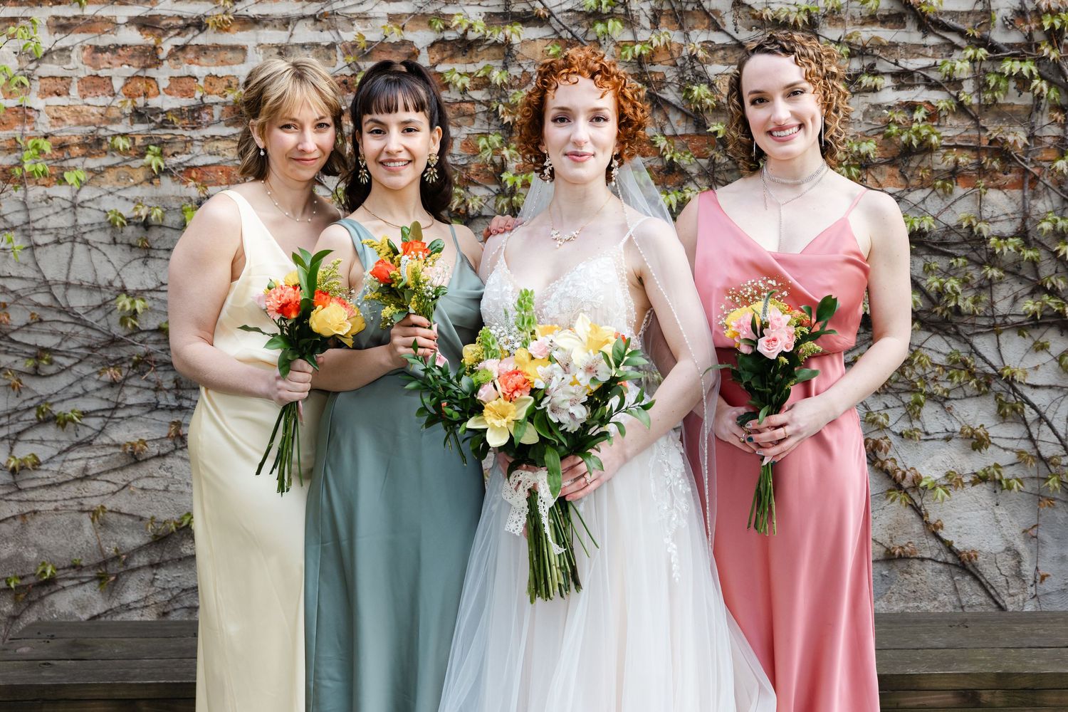 Bride in white gown with three bridesmaids in colorful mismatched dresses holding bouquets against brick wall.