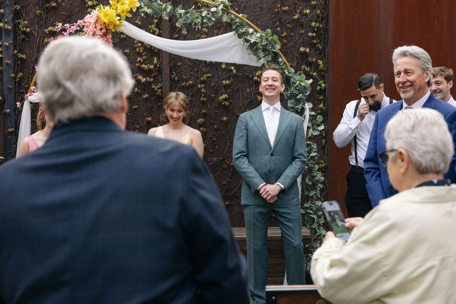 Groom in gray suit smiles at altar under floral arch during outdoor wedding ceremony with guests watching.