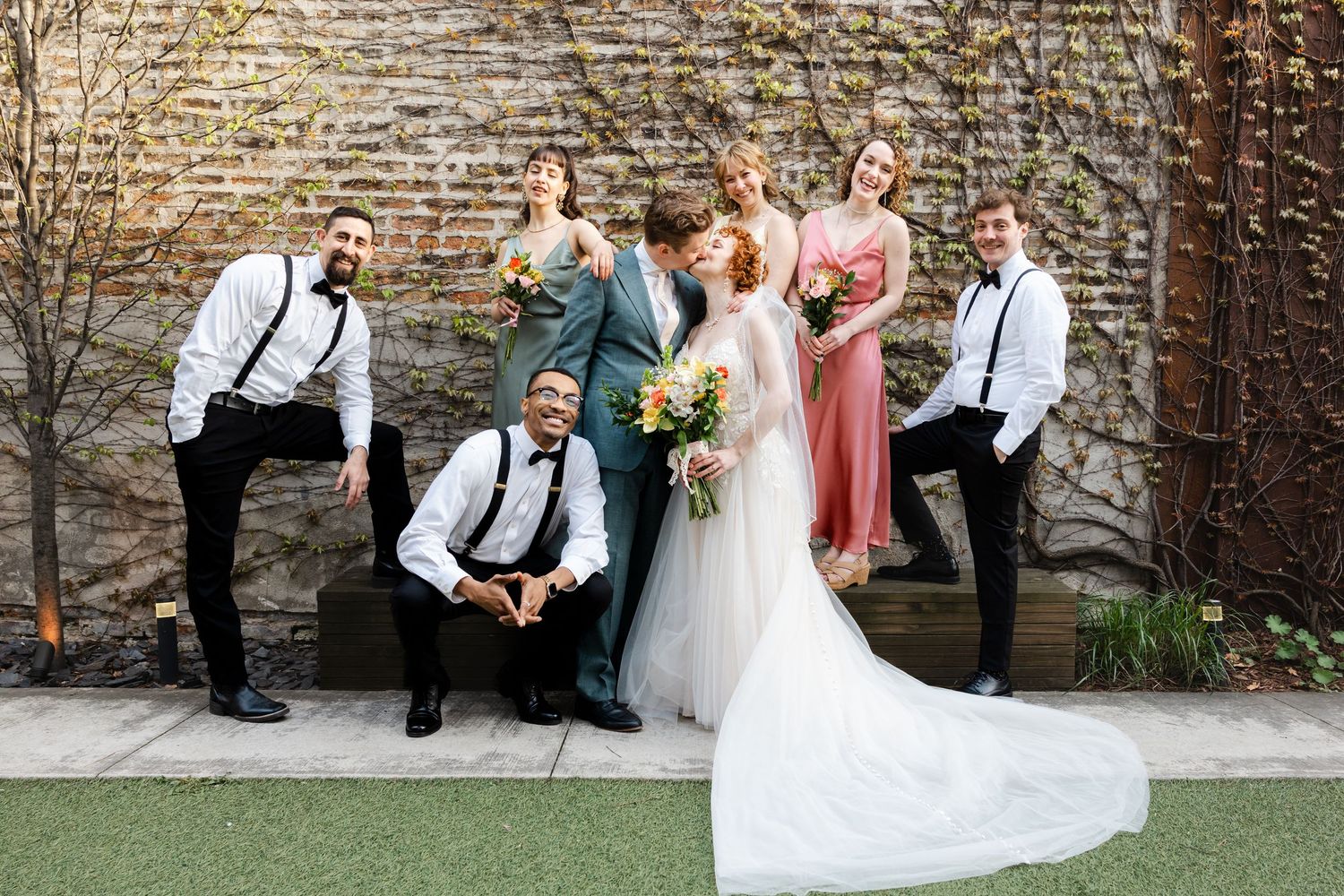 Wedding party poses playfully in front of a brick wall with ivy, bridesmaids in colorful dresses and groomsmen in suspenders.