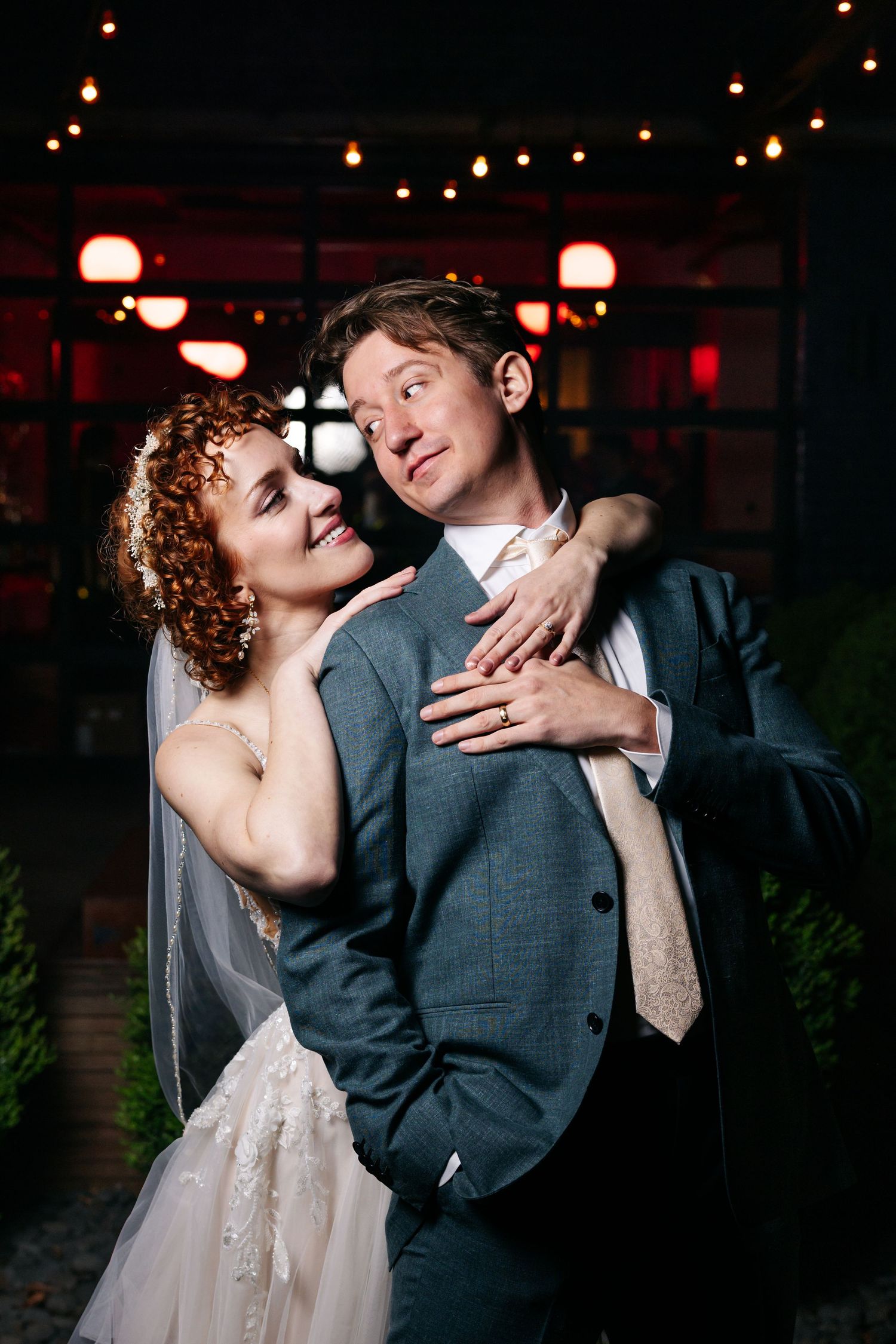Bride with red curly hair embraces her groom in a gray suit under string lights at their evening reception.