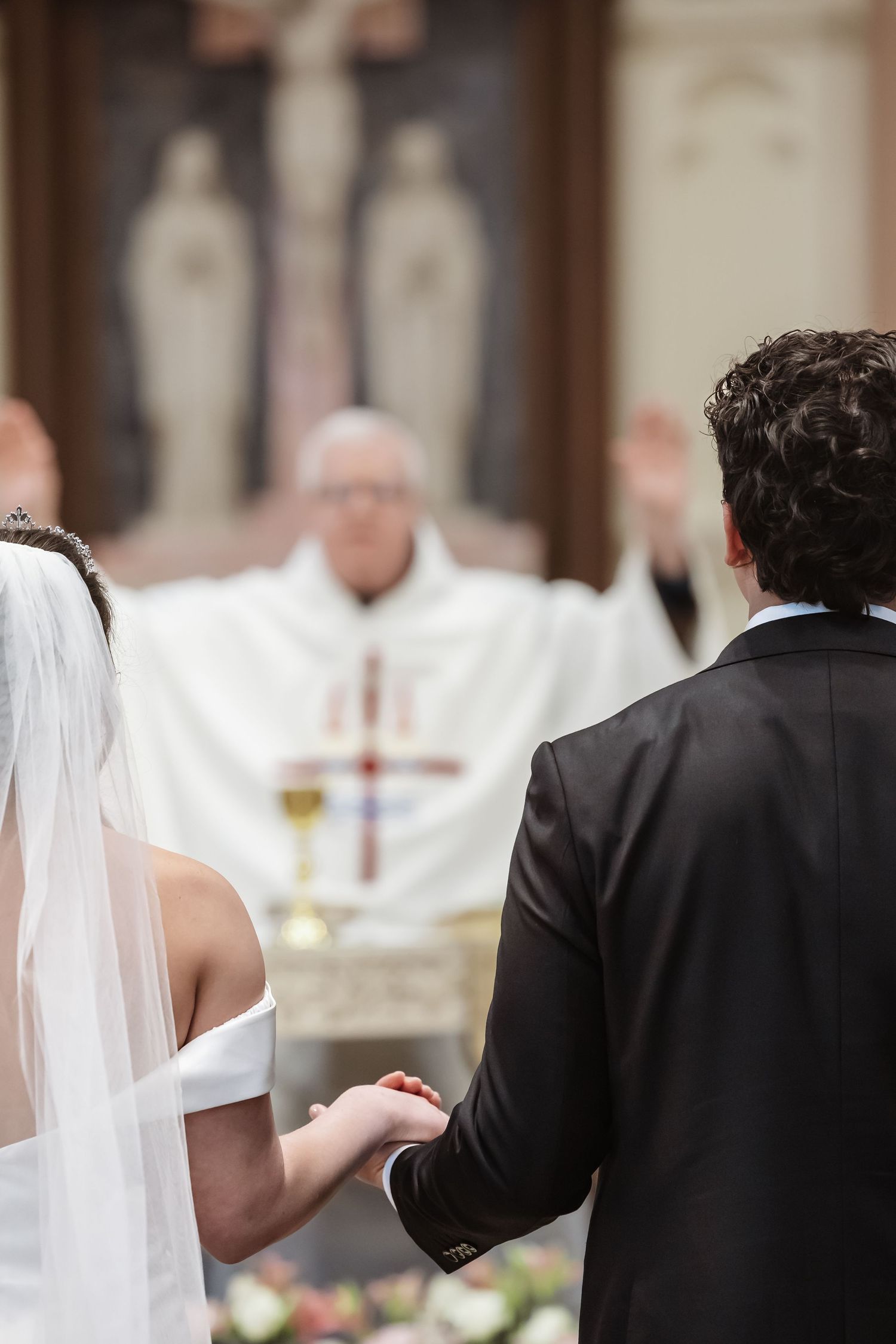 A bride and groom hold hands at the altar during a Catholic wedding ceremony with a priest blessing them.