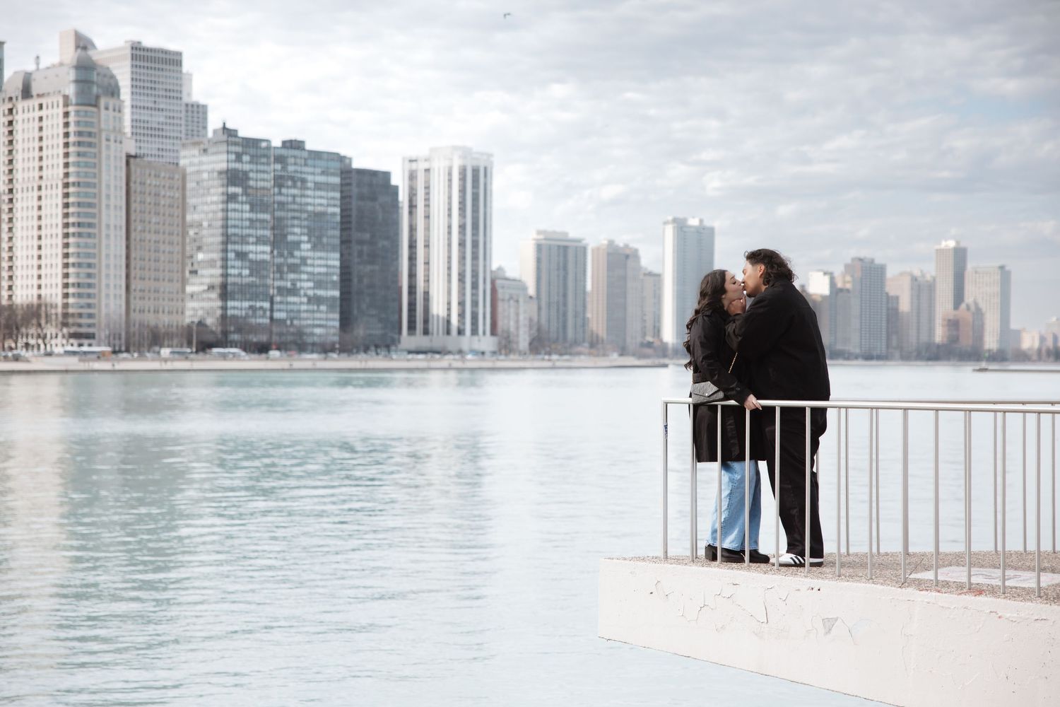Couple kissing by a railing with the Chicago skyline and lake reflecting in the background.