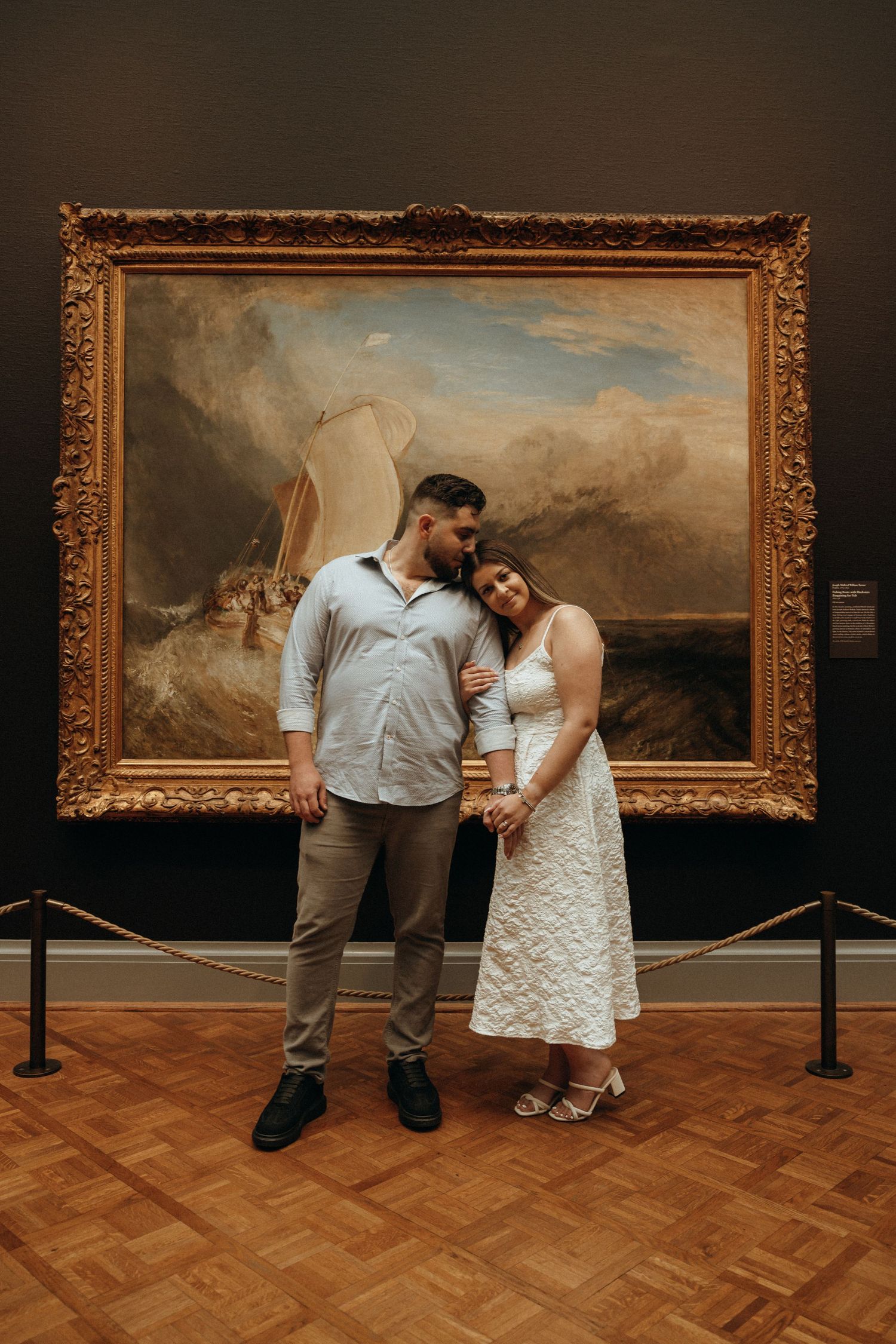 Couple posing tenderly before a grand Turner seascape painting in a golden frame at an art museum.