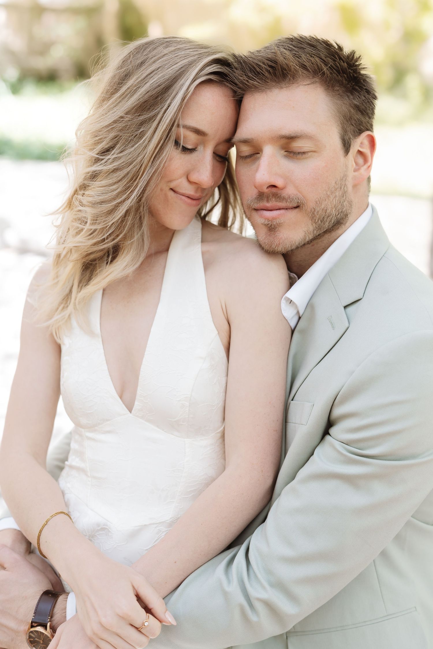 Groom in sage suit tenderly embraces bride with eyes closed in a bright outdoor portrait.