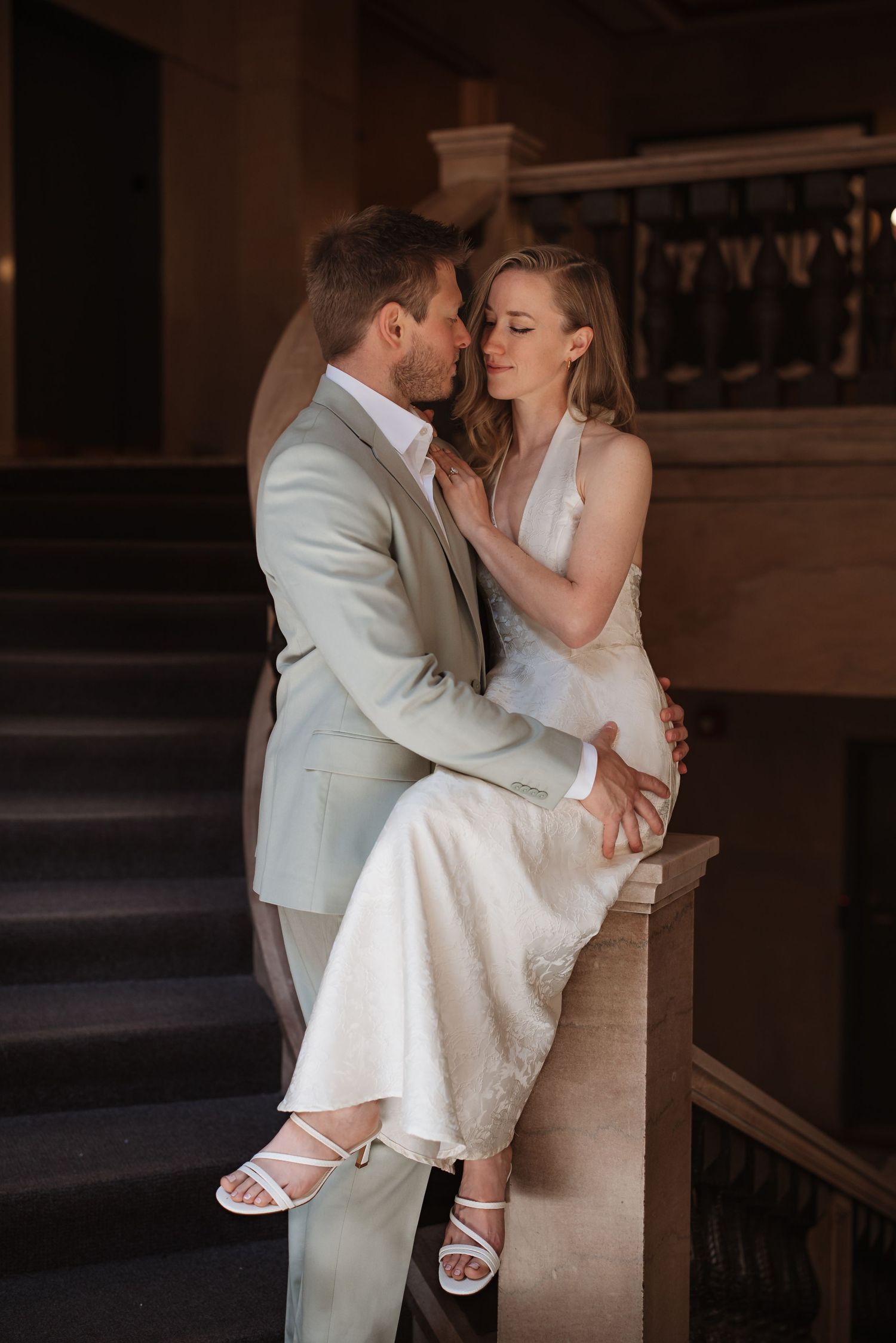Groom embraces seated bride on ornate staircase railing in elegant indoor wedding photo.