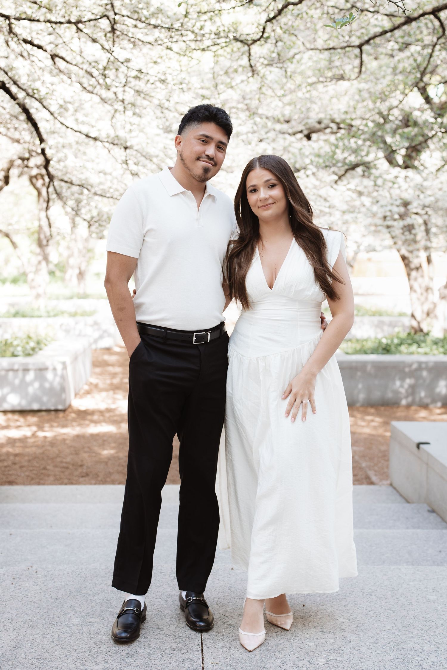Couple posing outdoors under blooming white trees, man in white polo and black pants, woman in white dress.