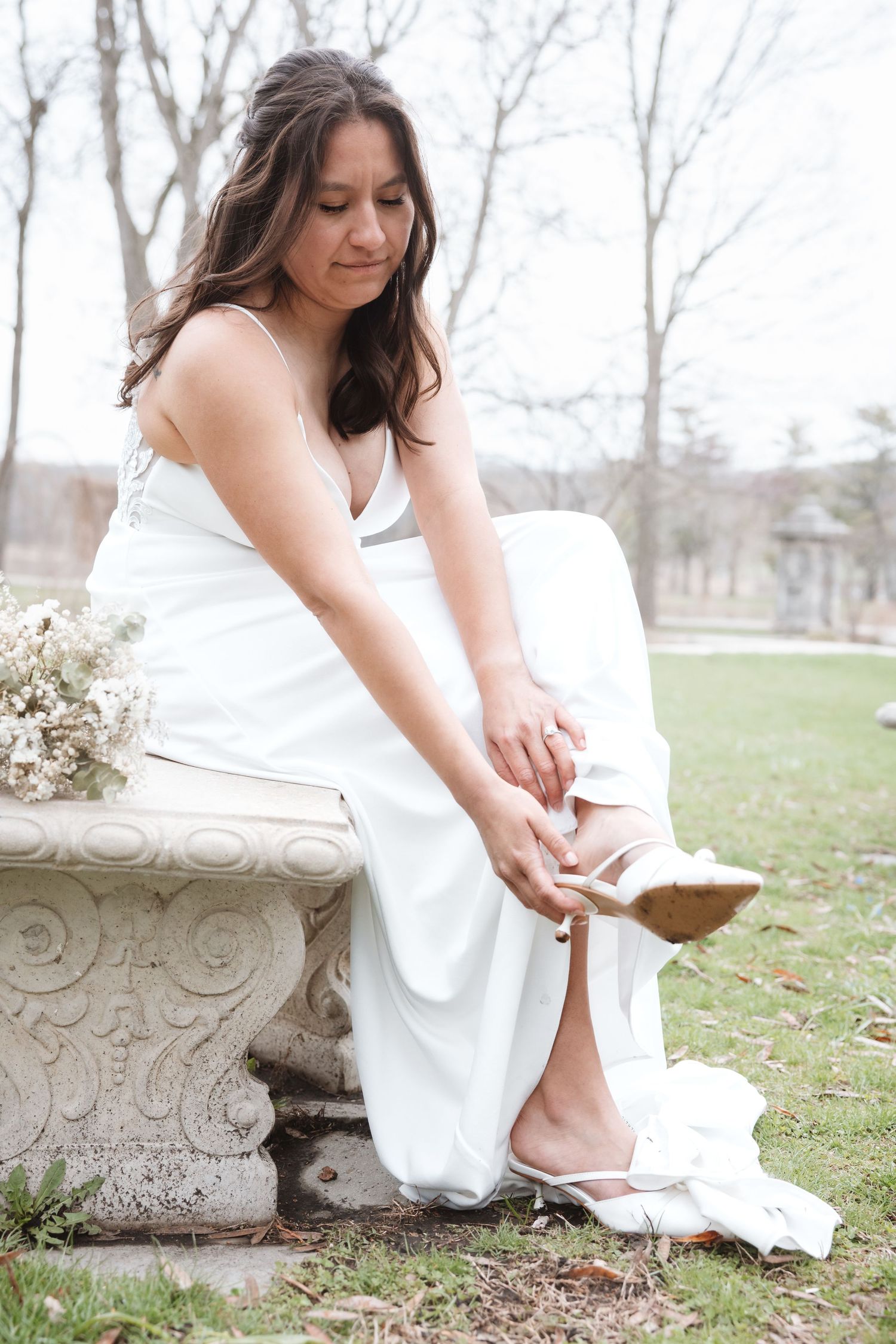 Bride in white gown sitting on stone bench adjusting her heels outdoors with bouquet beside her.