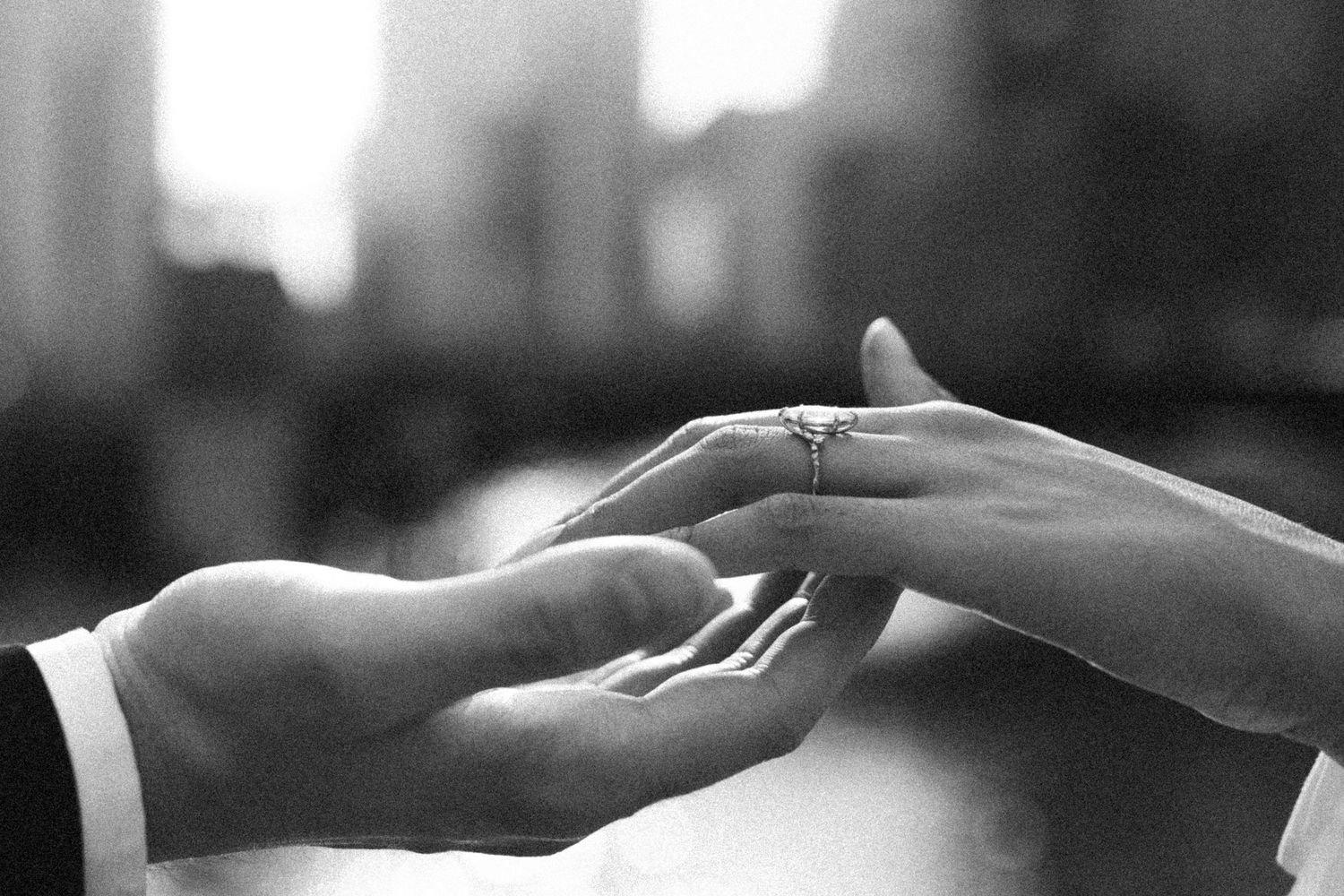 Black and white close-up of groom placing diamond engagement ring on bride's hand in the city.