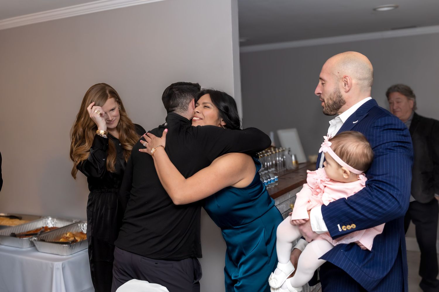 Wedding guests celebrate and hug while man in blue suit holds newborn baby at reception.