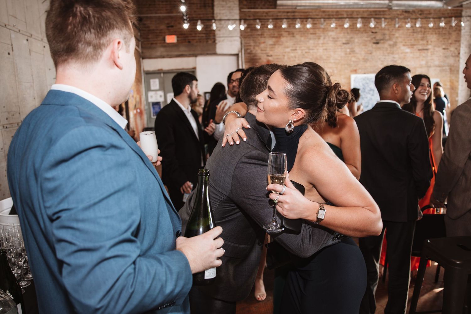 Woman holding champagne embraces a guest at an elegant indoor cocktail party.