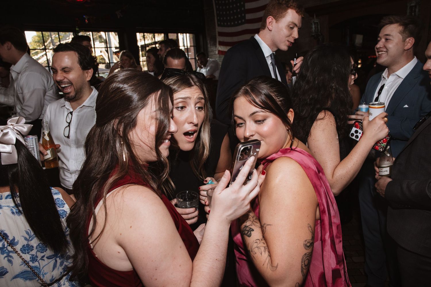 Three women in formal dresses laugh and take photos together at a festive wedding reception.