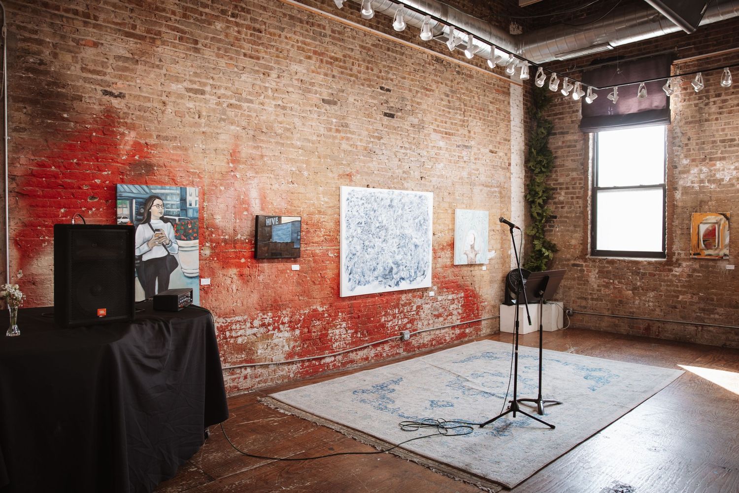 Art gallery loft space with exposed brick walls displaying paintings, a microphone stand, and natural window light.
