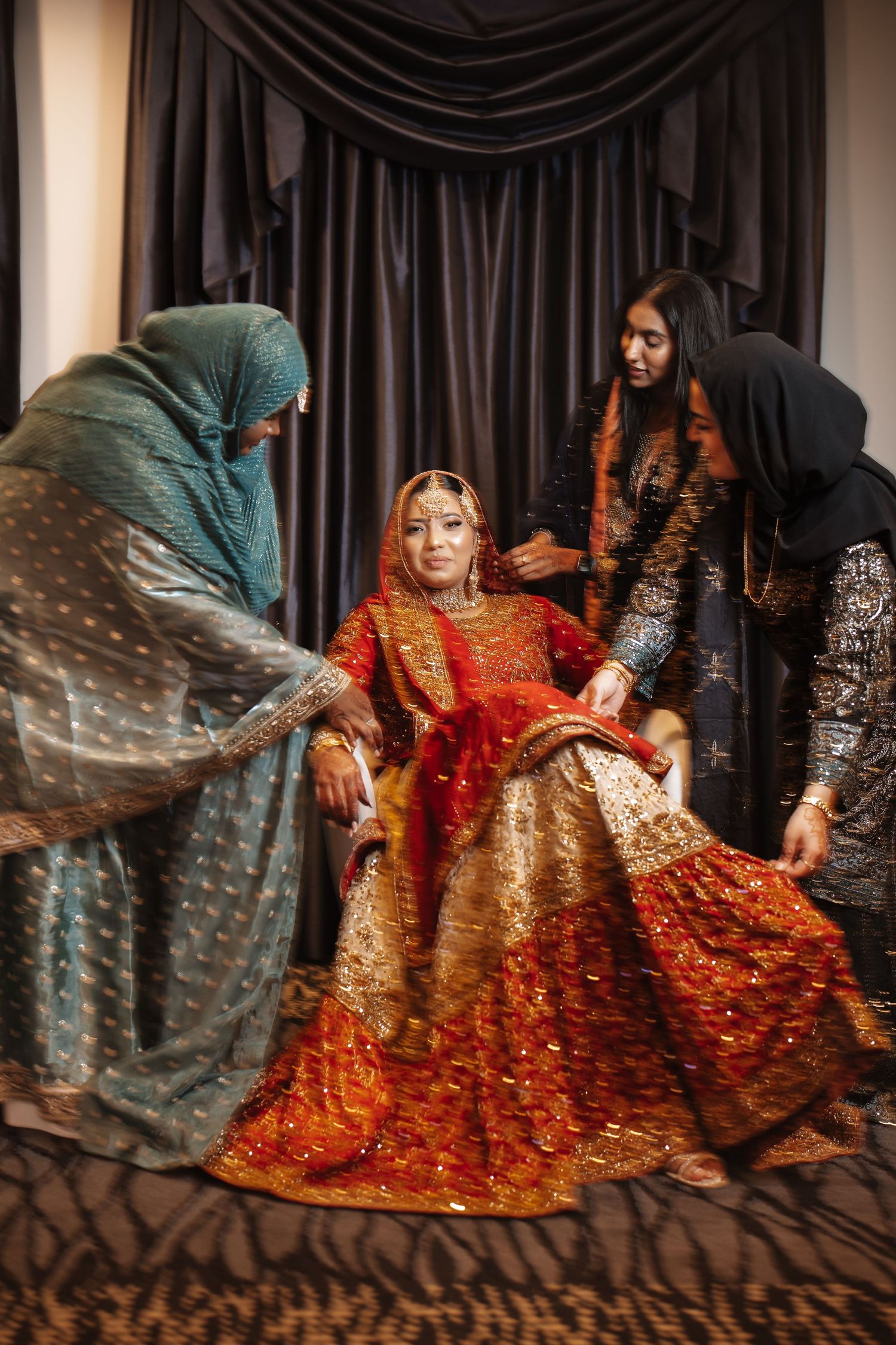 South Asian bride in embroidered red lehenga seated while attendants in hijabs adjust her outfit.