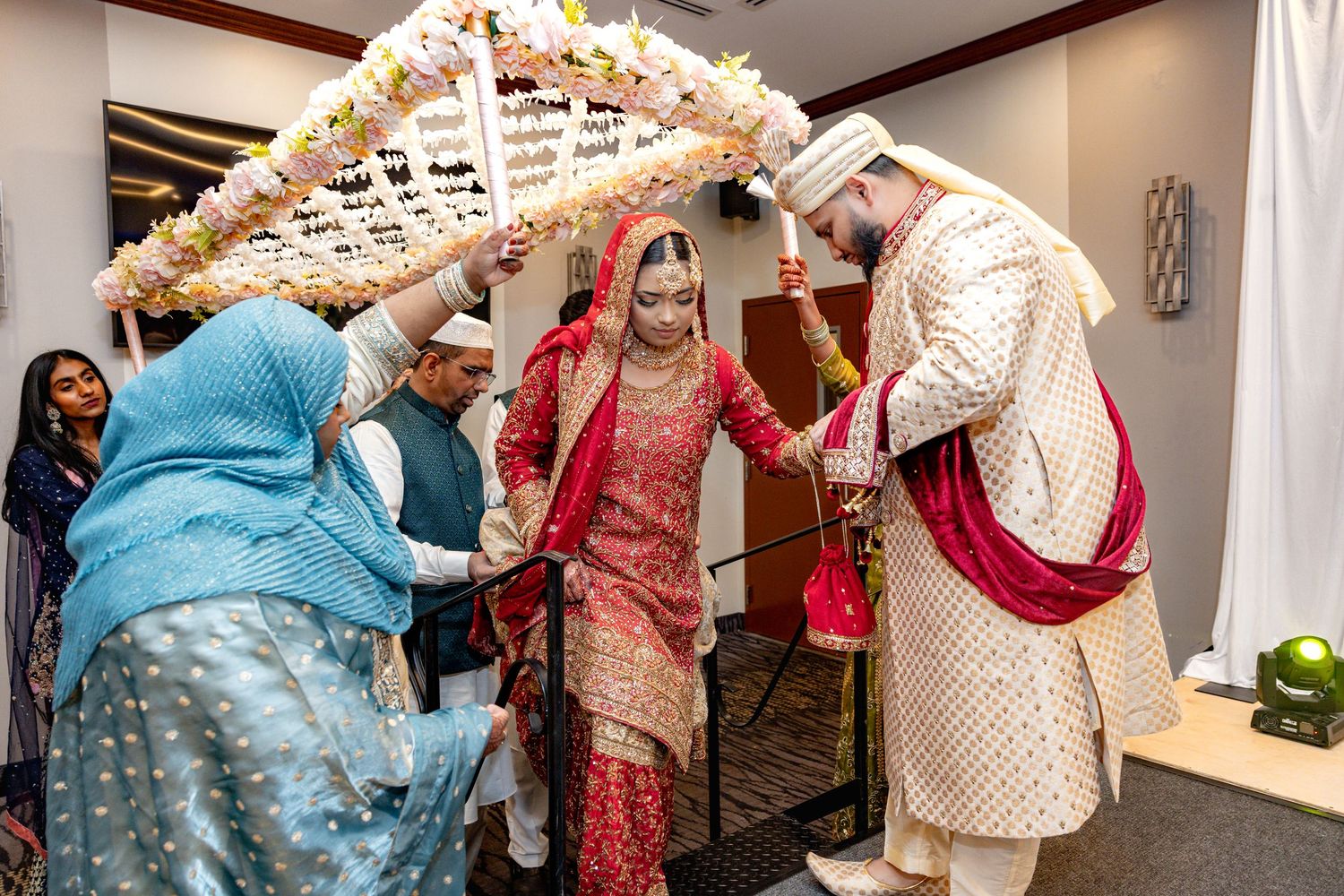 South Asian wedding couple walking under decorative floral arch assisted by family members indoors.