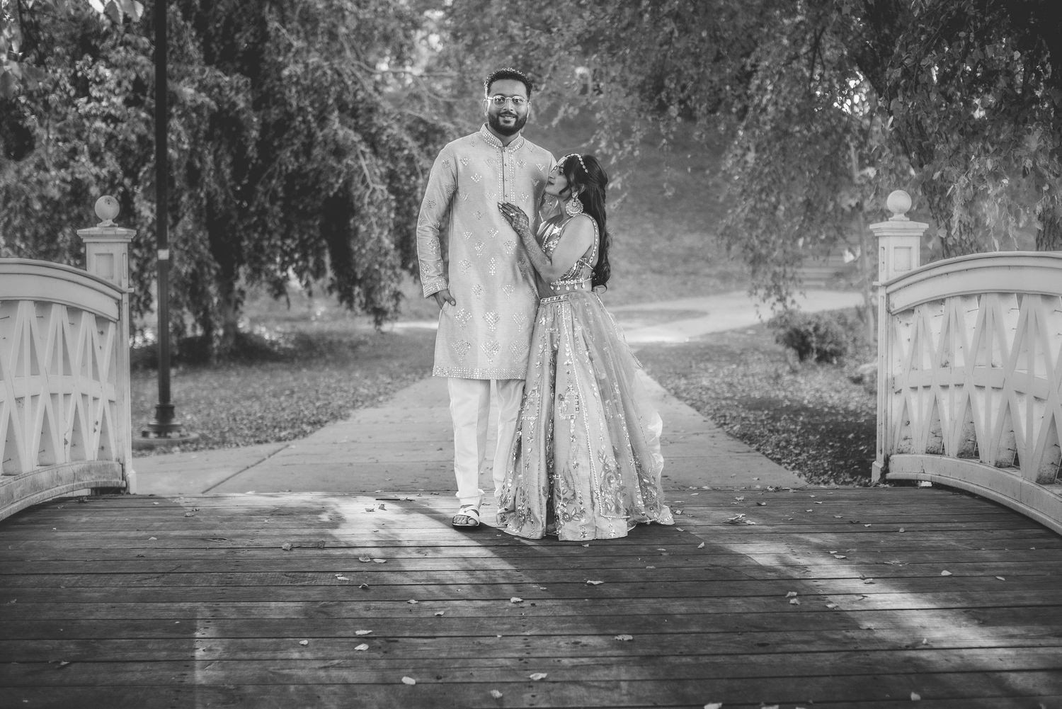 Black and white photo of Indian couple in traditional attire standing on wooden bridge.