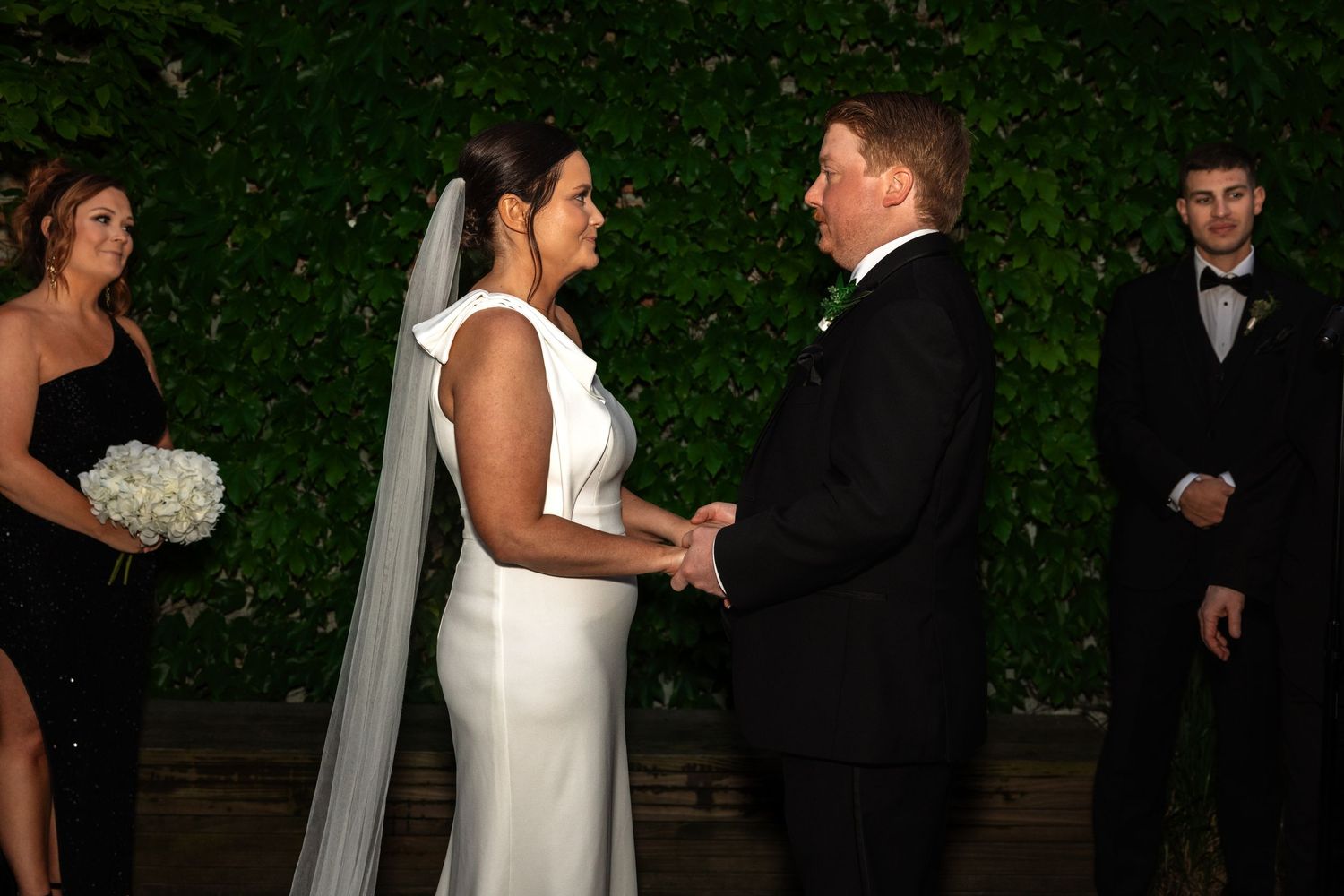 Bride and groom hold hands during outdoor evening wedding ceremony with ivy wall backdrop.
