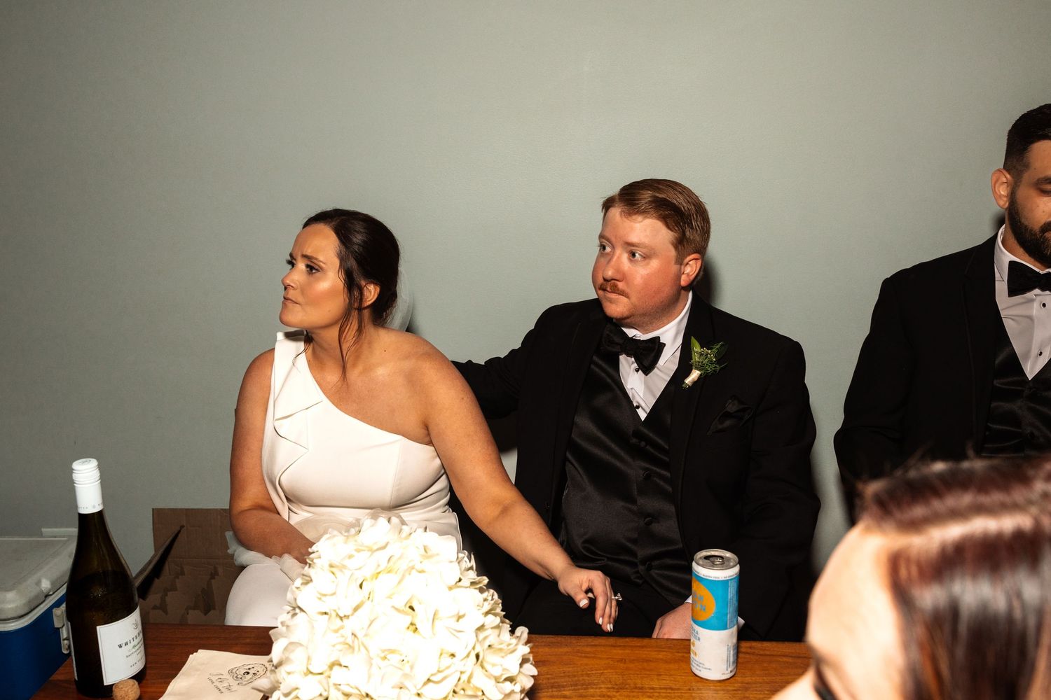 Bride in white one-shoulder gown and groom in tuxedo seated at wedding reception table with flowers.