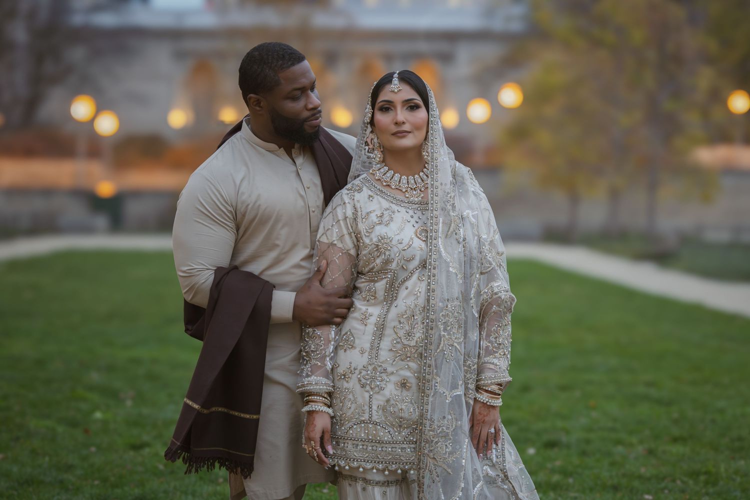 Interracial Muslim couple in traditional South Asian bridal outfits sharing intimate moment in park at dusk.