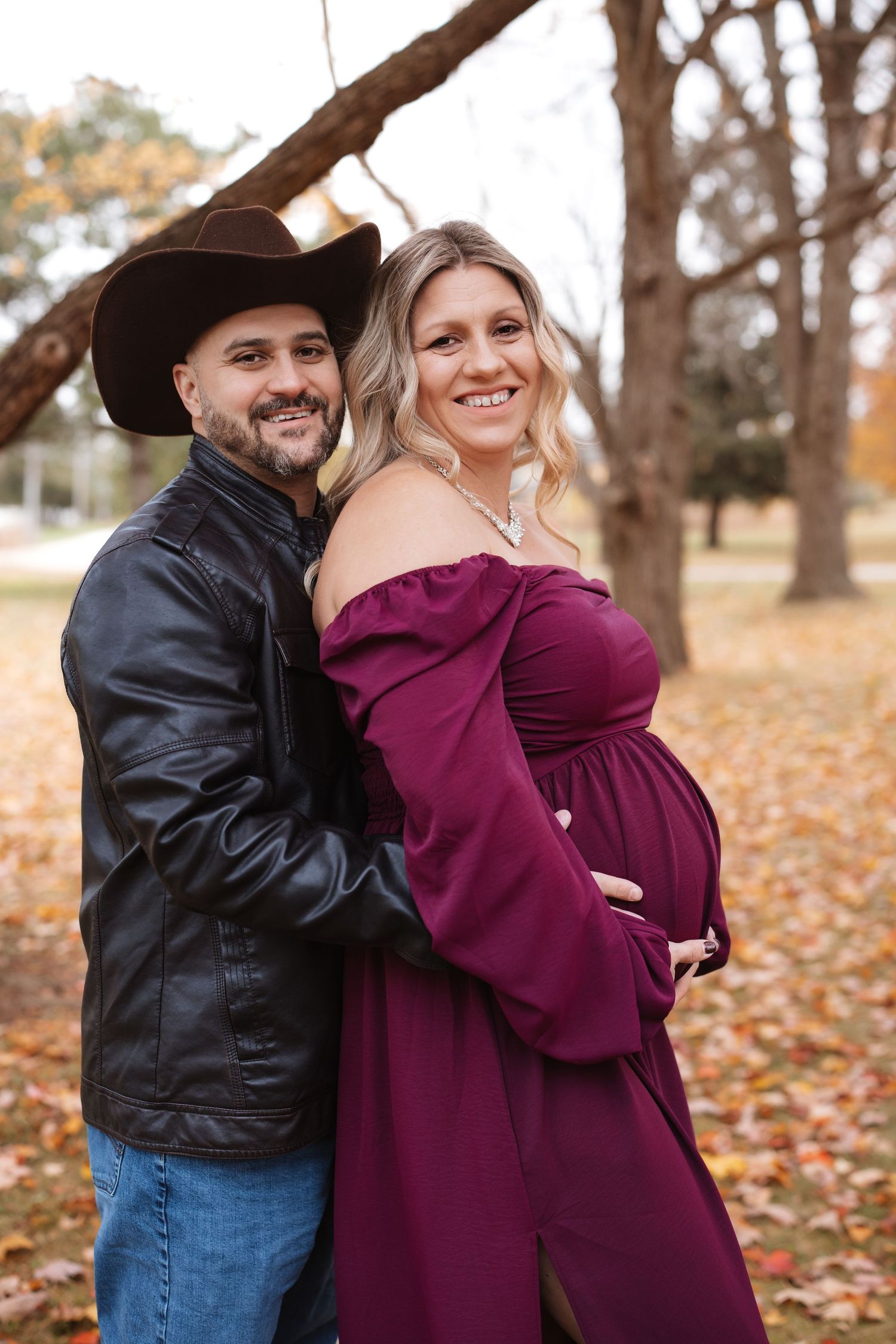Happy couple maternity portrait outdoors, man in cowboy hat and leather jacket with pregnant woman in burgundy dress.