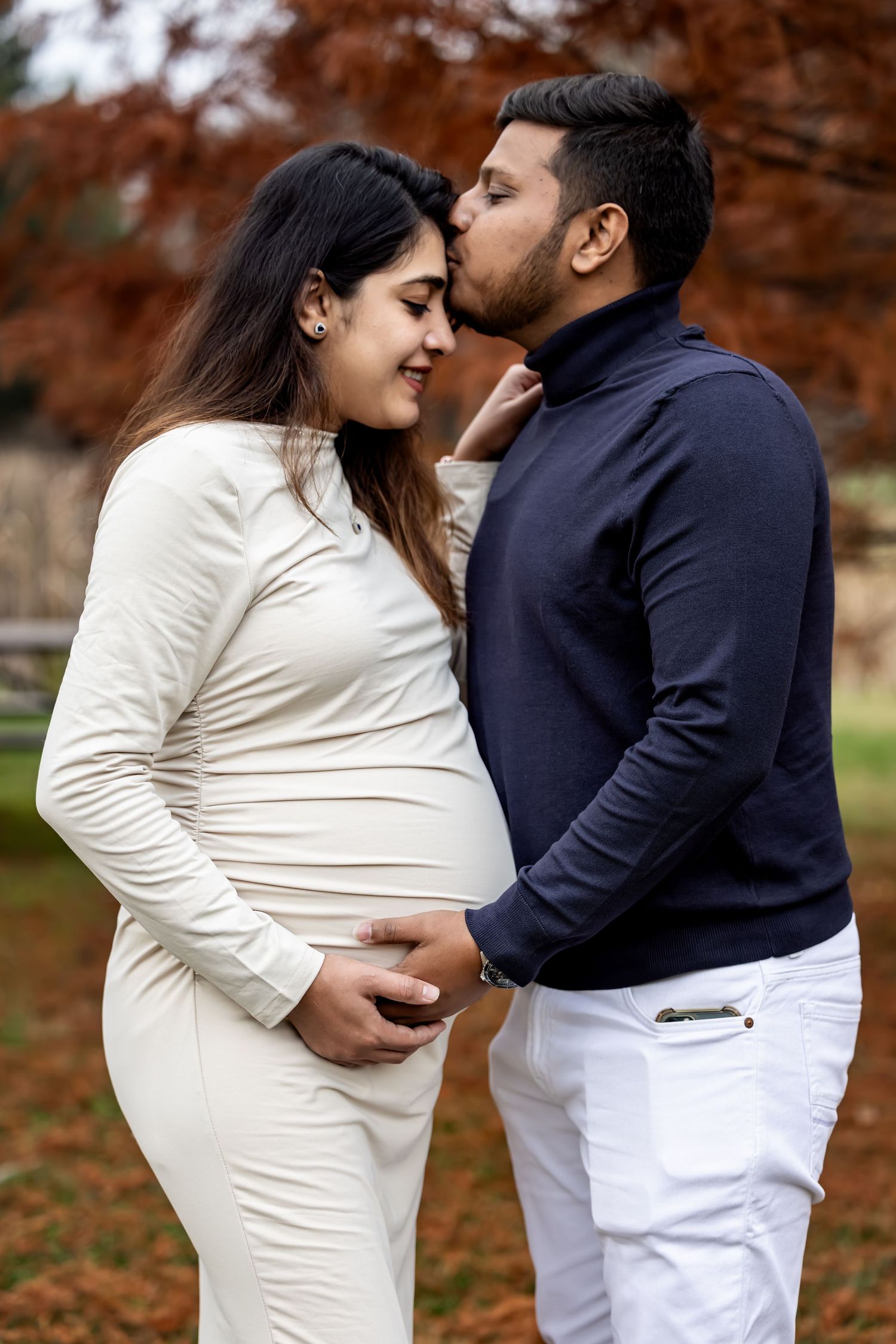 Man in navy sweater kissing pregnant woman's cheek while holding her baby bump in autumn park.