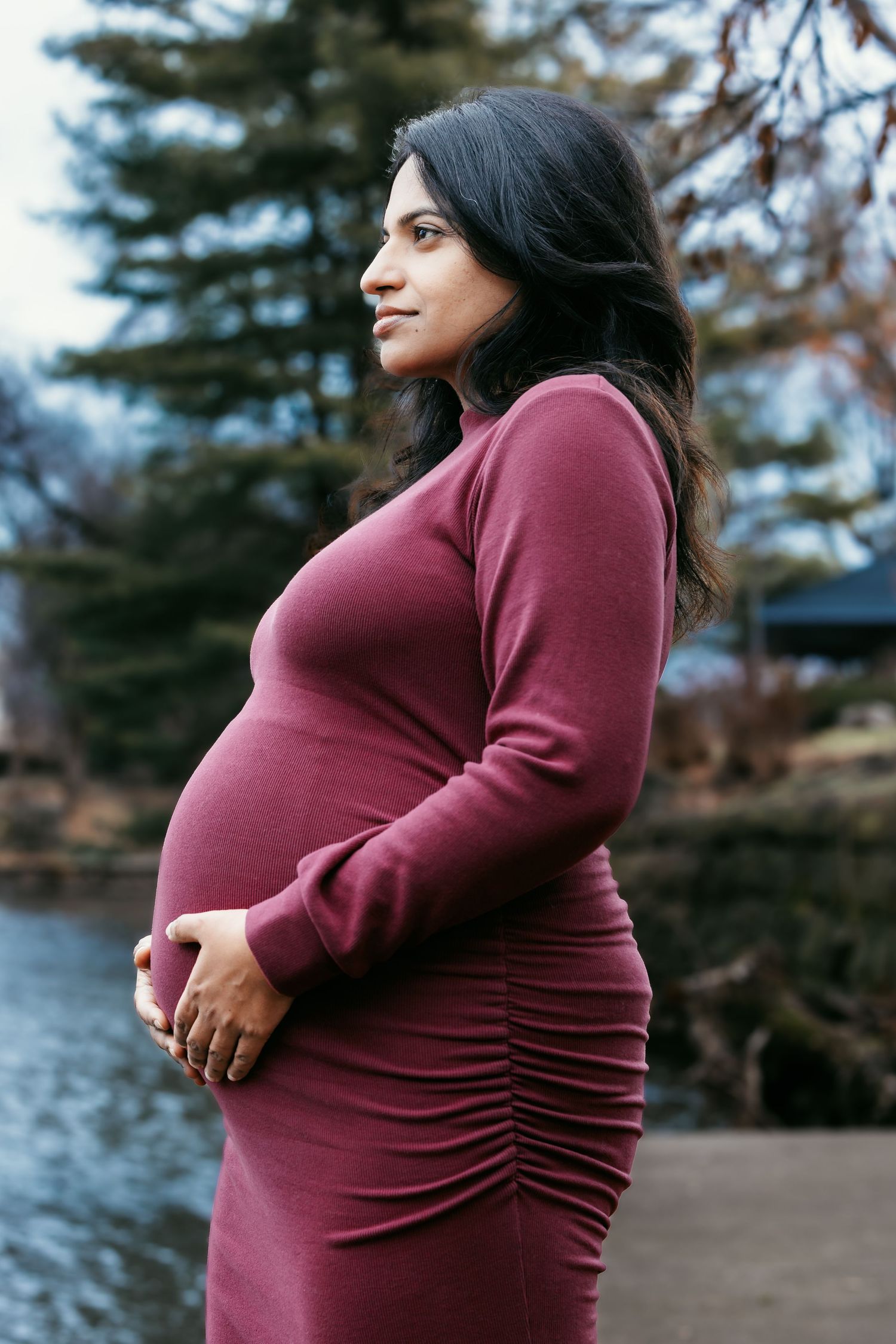 Pregnant woman in fitted burgundy dress posing sideways, holding belly near a tranquil park pond.