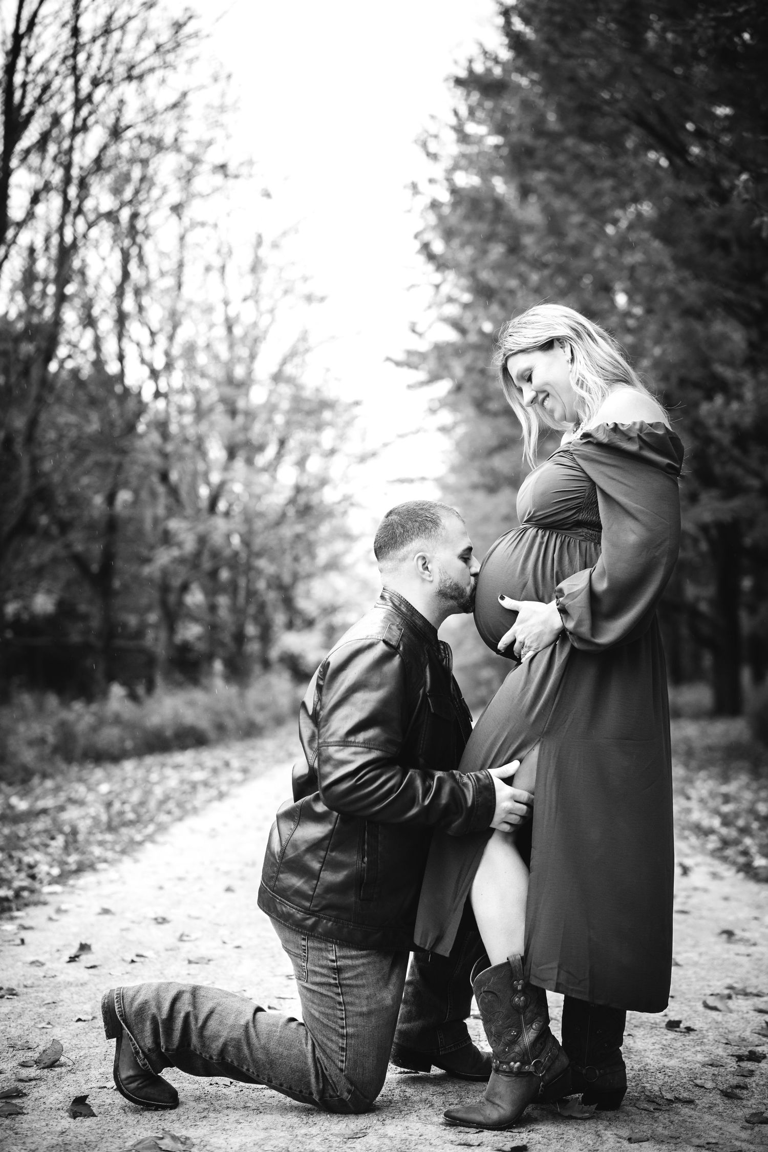 Black and white photo of a man kneeling and kissing pregnant woman's belly on a forest path in autumn.