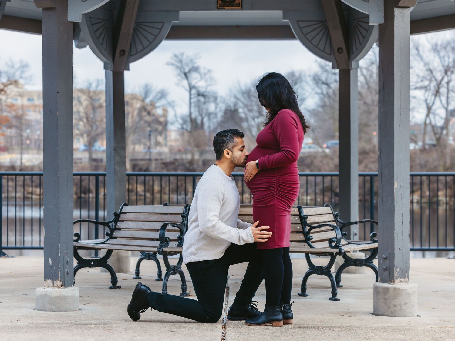 Man on one knee holding hands with pregnant woman in burgundy dress under a park gazebo.