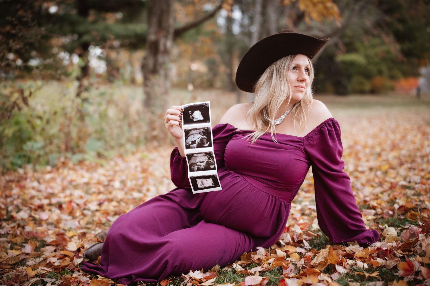Pregnant woman in purple dress and black hat sitting in fall leaves holding ultrasound photos outdoors.